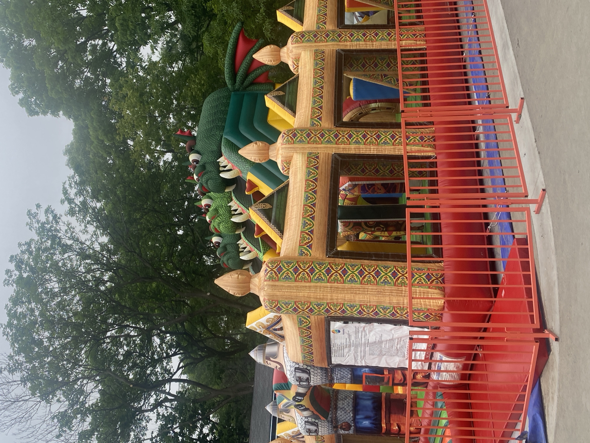 An inflatable dragon-themed bouncy castle is set up outdoors, surrounded by a red metal fence. The bouncy castle features multiple green dragon heads with red horns and large red eyes across the top, along with ornate, wood-like patterned walls and decorative finials. Behind the bouncy castle, lush green trees and an overcast sky are visible. The ground in front of the bouncy castle is covered with red safety mats and blue tarps. No people are visible in the image. The scene appears to be in a park or outdoor event space.