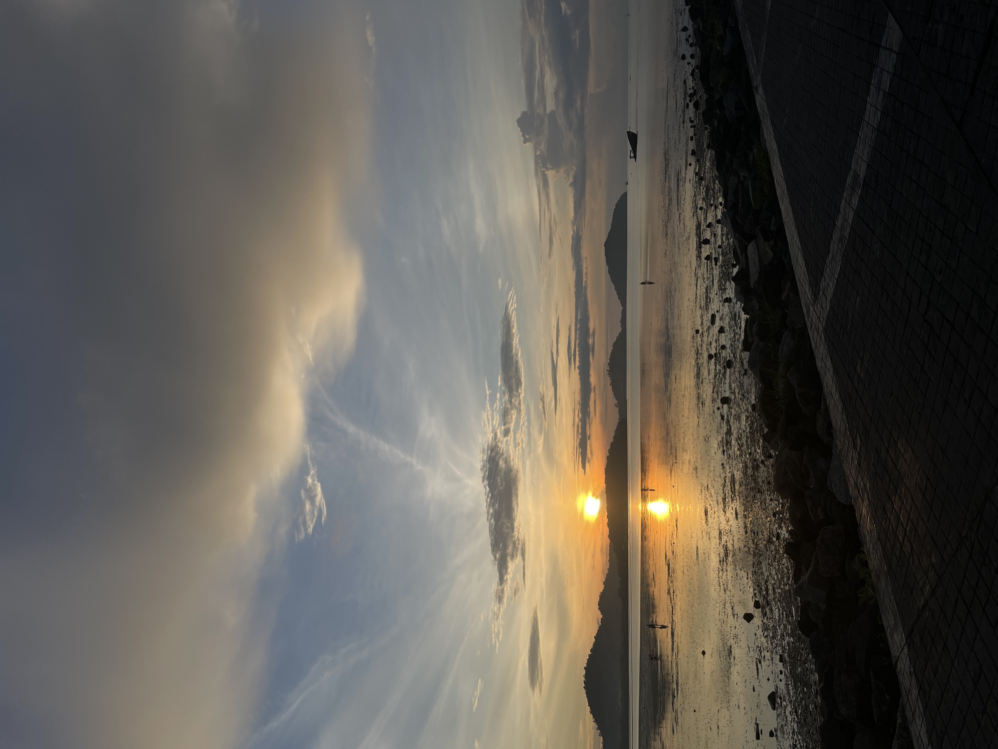 This is a photograph of a sunset over the ocean in Phuket City Municipality, Thailand. The sun is setting behind a series of islands in the distance, casting a golden glow on the water. The sky is filled with clouds in shades of gray and orange, with some wispy clouds also visible. In the foreground, there is a paved walkway along the shore, with rocks and seaweed scattered on the beach. There are a few people visible in the distance, wading in the shallow water. A small boat is also visible anchored in the ocean. The overall mood of the photograph is peaceful and serene, capturing the beauty of a tropical sunset.