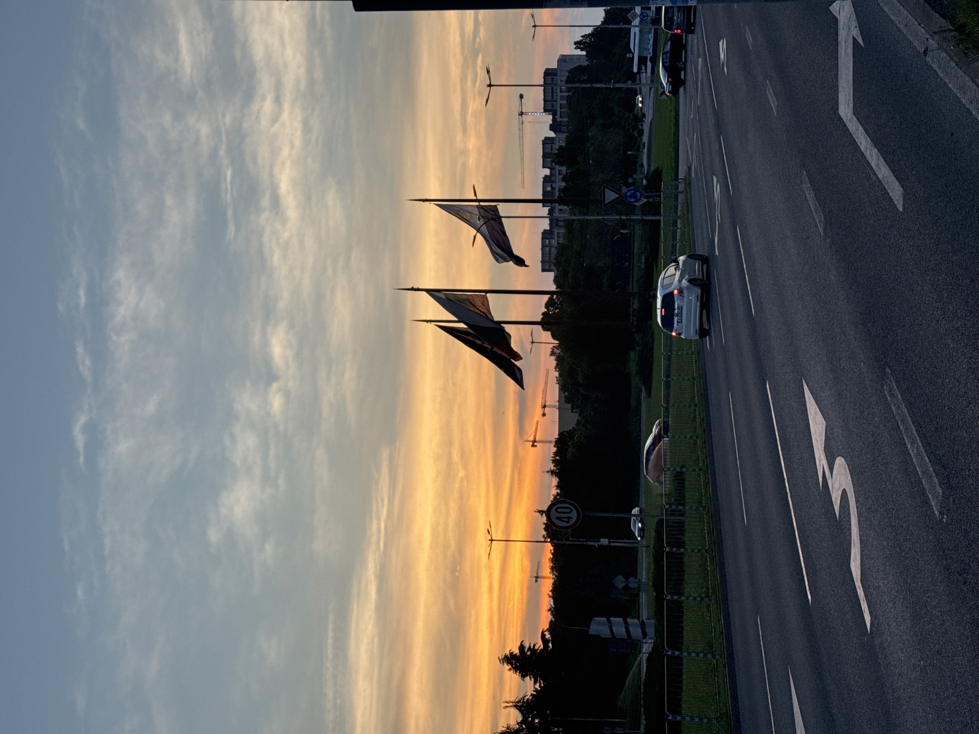 The image depicts an urban landscape at dusk in the city of Unknown, Unknown. A wide, multi-lane asphalt road with prominent white lane markings, including a large, curved left-turn arrow in the foreground, dominates the lower half of the frame. Several vehicles are visible: a white hatchback in the middle distance with illuminated taillights, a dark-colored sedan, and an orange/red car on an adjacent lane.
A green median separates traffic lanes, featuring a metal fence, multiple streetlights, and traffic signs, including a circular "40" speed limit sign and a triangular yield sign. Three flags on tall poles are erected on the median, their designs partially discernible against the twilight sky. The flags appear to be tricolors, with one potentially showing a coat of arms.
The sky transitions from a vivid orange and yellow sunset along the horizon to a lighter blue with scattered wispy clouds in the upper portion. Silhouetted trees and dark buildings form the distant skyline, interspersed with at least three large construction cranes, suggesting ongoing development. No individuals are clearly visible. The overall scene conveys an active urban environment transitioning from day to night.