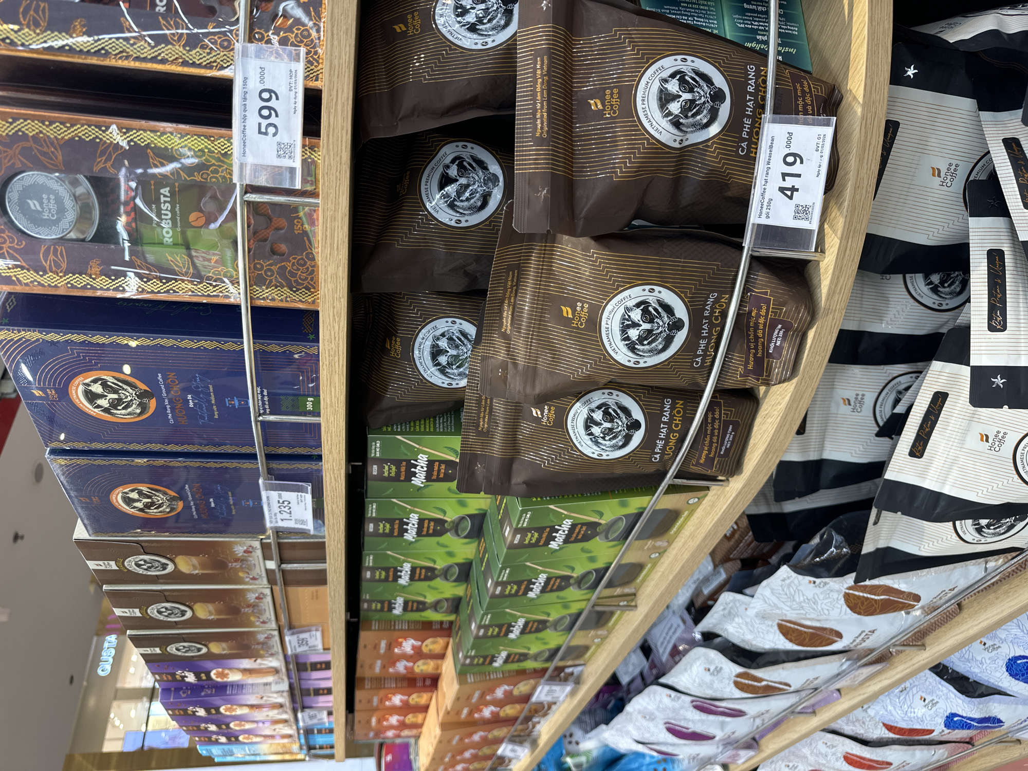 This media file is a close-up, eye-level shot of a retail display shelf filled with coffee and matcha products. The shelf is made of light-colored wood and appears to be in a well-lit store, possibly a supermarket or specialty food shop. The products are arranged in neat rows and stacked on top of each other.
Dominating the foreground are several packages of "Honee Coffee," which feature an illustration of an animal, likely a civet, and Vietnamese text that includes "CÀ PHÊ HẠT RANG HƯƠNG CHỒN." Some of these packages have price tags, one indicating "419.000₫" and another "599.000₫," along with details like "gói 250g" and "hộp quà tặng 150g." Other coffee products include those labeled "ROBUSTA" and "BAN ME GOLD."
Interspersed with the coffee are boxes of "Matcha" products, with one visible price tag of "1.235.000₫." The matcha boxes are primarily green and feature imagery of matcha tea bowls.
In the background, more coffee packages are visible, some with a black and white design. The overall impression is one of abundance and variety in coffee and tea offerings. The lighting suggests it's daytime, and the setting is an indoor retail environment in Vietnam, as indicated by the Vietnamese text on the products and price tags. There are no people visible in the frame.