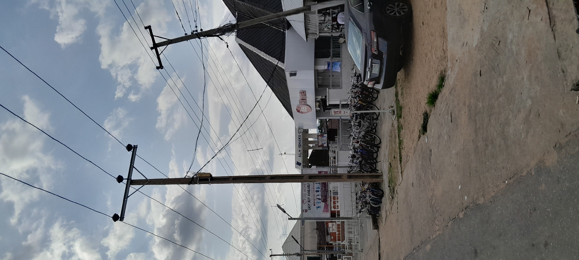 In Jalingo, Nigeria, a street scene unfolds under a cloudy sky. A large wooden utility pole dominates the foreground, adorned with a transformer and a tangle of overhead wires. To the left, a white building with a triangular roof features a prominent banner advertising "ITEL HOME EASY BUY AVAILABLE." Further to the right, another building, also white, has the "itel Home" logo displayed. In front of these establishments, a line of parked bicycles suggests a busy commercial area. A dark-colored sedan is parked to the right, its rear end visible. The ground is a mix of paved and unpaved surfaces, with patches of dry grass. The overall impression is one of an active, perhaps bustling, urban environment.