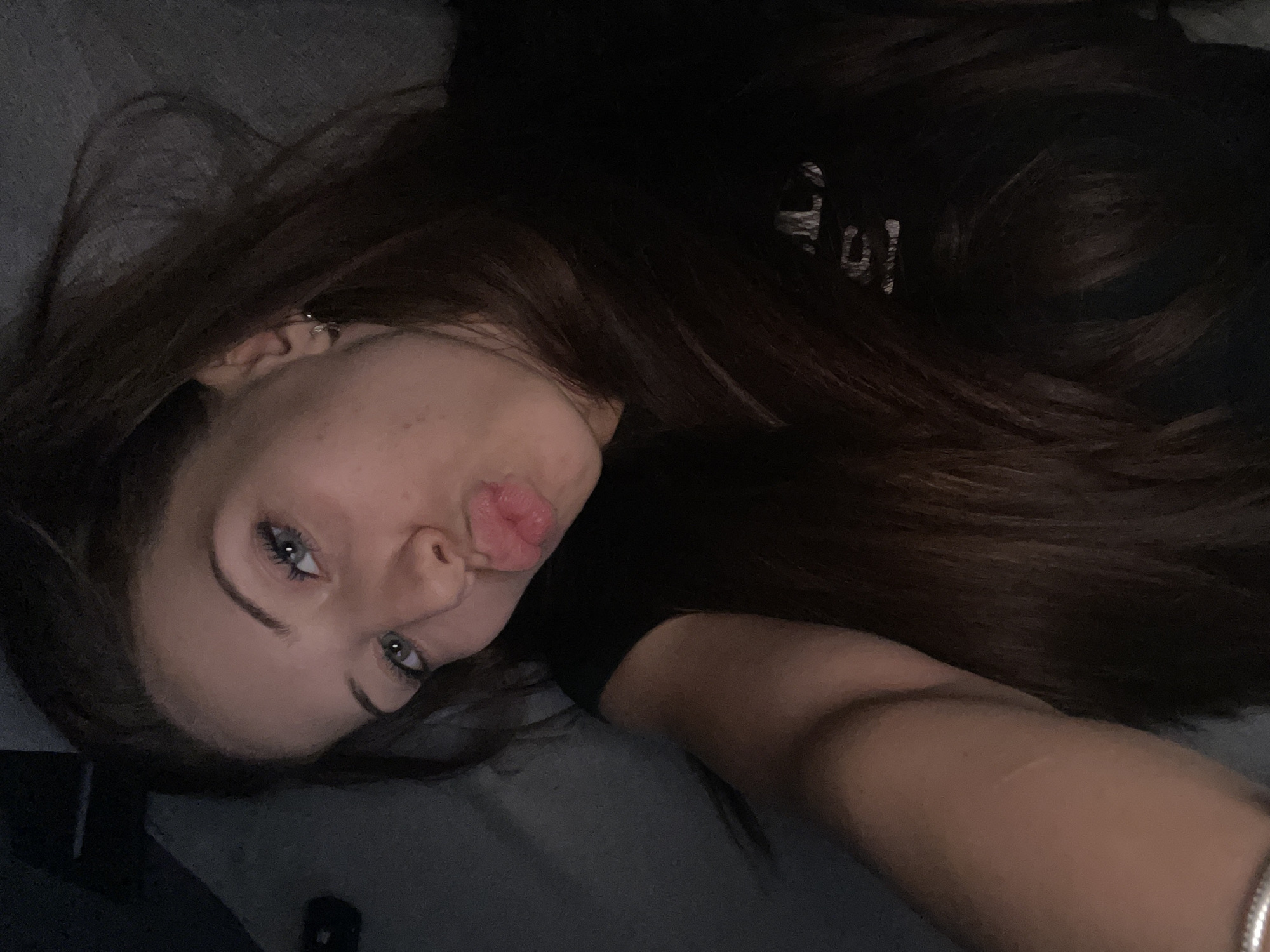In a dimly lit indoor setting, a young woman with long, dark brown hair is captured in a close-up, self-taken photograph. Her head is tilted, and her body is angled towards the camera, suggesting she is lying down or reclined. She has striking blue-green eyes and is making a "duck face" with her lips. She is wearing a black t-shirt, and a glimpse of a silver bracelet can be seen on her left wrist. The background is dark and appears to be a textured surface, possibly a couch or bed, with a subtle gradient of light and shadow. The lighting is low, casting a soft glow on her face and highlighting her features. The overall impression is one of a casual, intimate moment captured in a personal space.