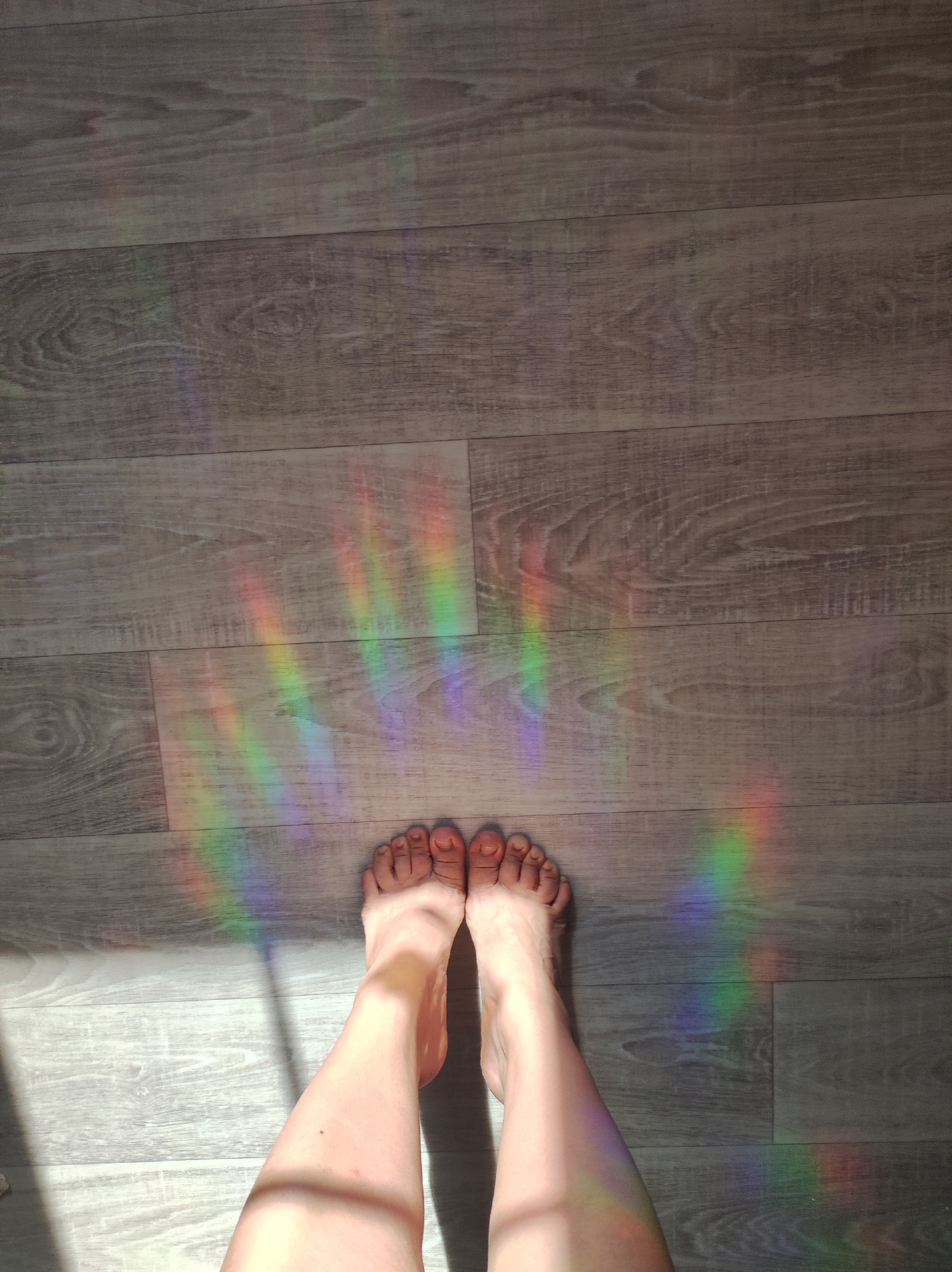 The image shows a first-person view of a person's legs and feet standing on a wooden floor. A rainbow refraction is cast on the floor in front of the person. The location is a room with wooden floors. No city is specified.