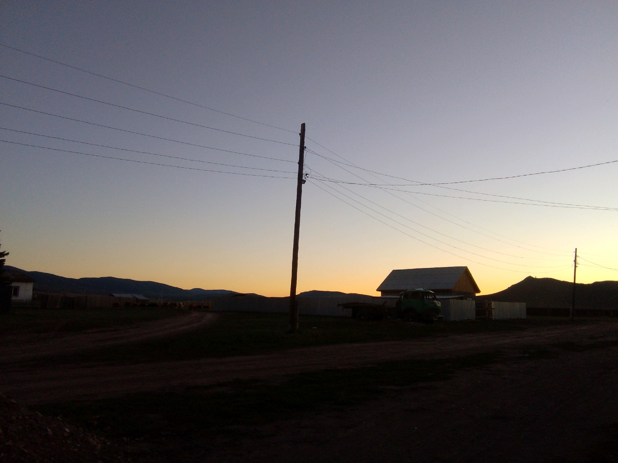 The image depicts a rural scene at dusk, possibly in or near the city of Sush, Russia, characterized by a silhouetted landscape against a gradient sky. A tall wooden utility pole stands prominently in the mid-ground, supporting multiple parallel power lines that stretch across the frame. In the background, a few low-lying buildings with angled roofs are visible, silhouetted against the fading light. A green truck is parked near one of the buildings. The foreground is dark and appears to be a dirt road or path. Hills or mountains form a dark, undulating line on the horizon. The sky transitions from a pale blue at the top to a warm yellow and orange hue near the horizon, indicating sunset or sunrise.
