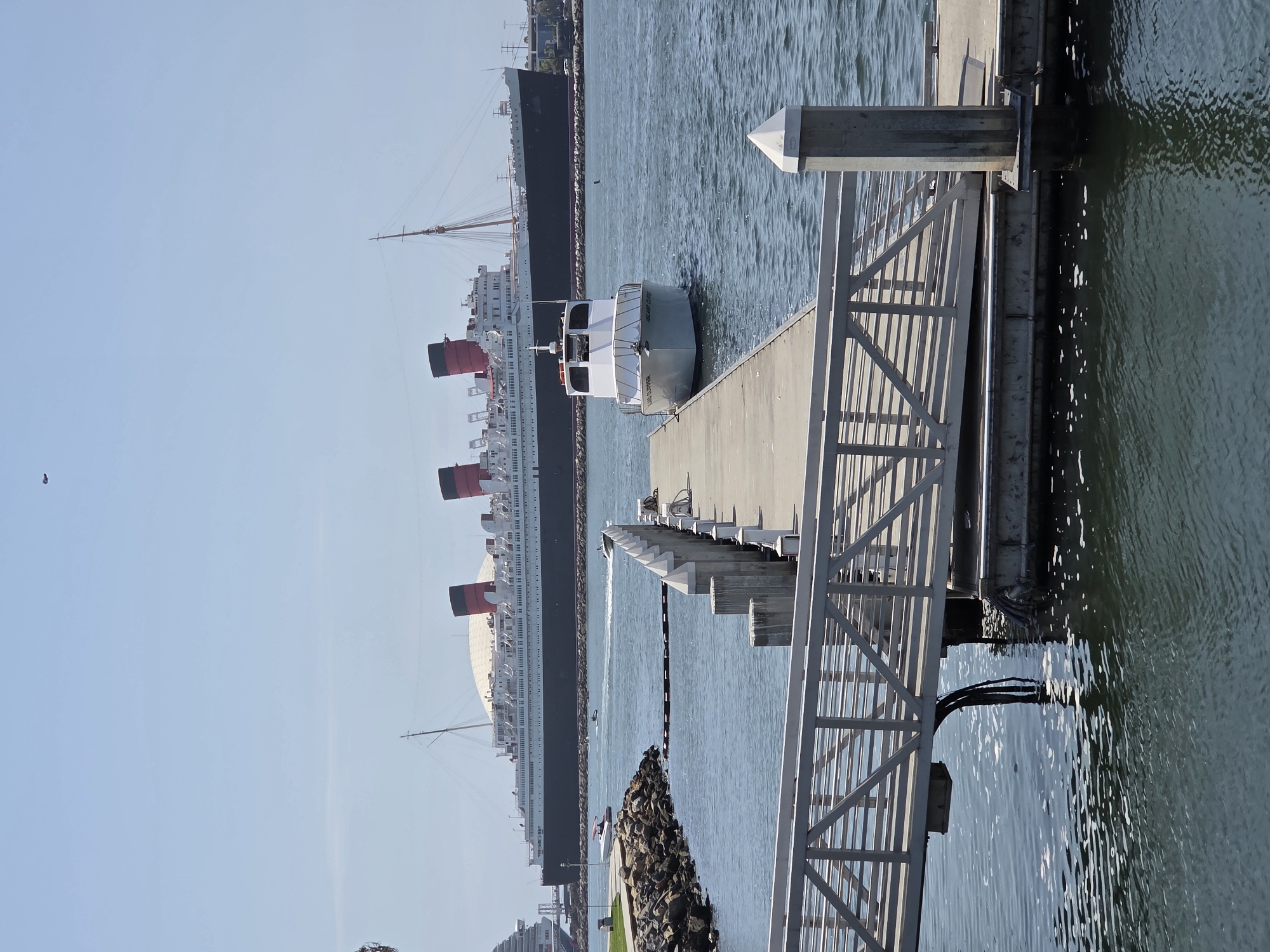 The image depicts a bright, sunny day at a harbor, likely near the Queen Mary ship in Long Beach, California. In the foreground, a concrete pier with a metal ramp extends into the water. Moored to the pier is a white boat with "ISLAND CLIPPER" visible on its side. Further in the background, the large ocean liner RMS Queen Mary is docked, its distinctive red and black funnels prominently displayed. The water is calm with gentle ripples, and a few smaller boats and a jet ski are visible in the distance. The sky is clear and blue, with a faint haze. The environment suggests a leisure or tourist area, with the Queen Mary serving as a major landmark. There are no people clearly visible in the immediate foreground or on the Queen Mary.
