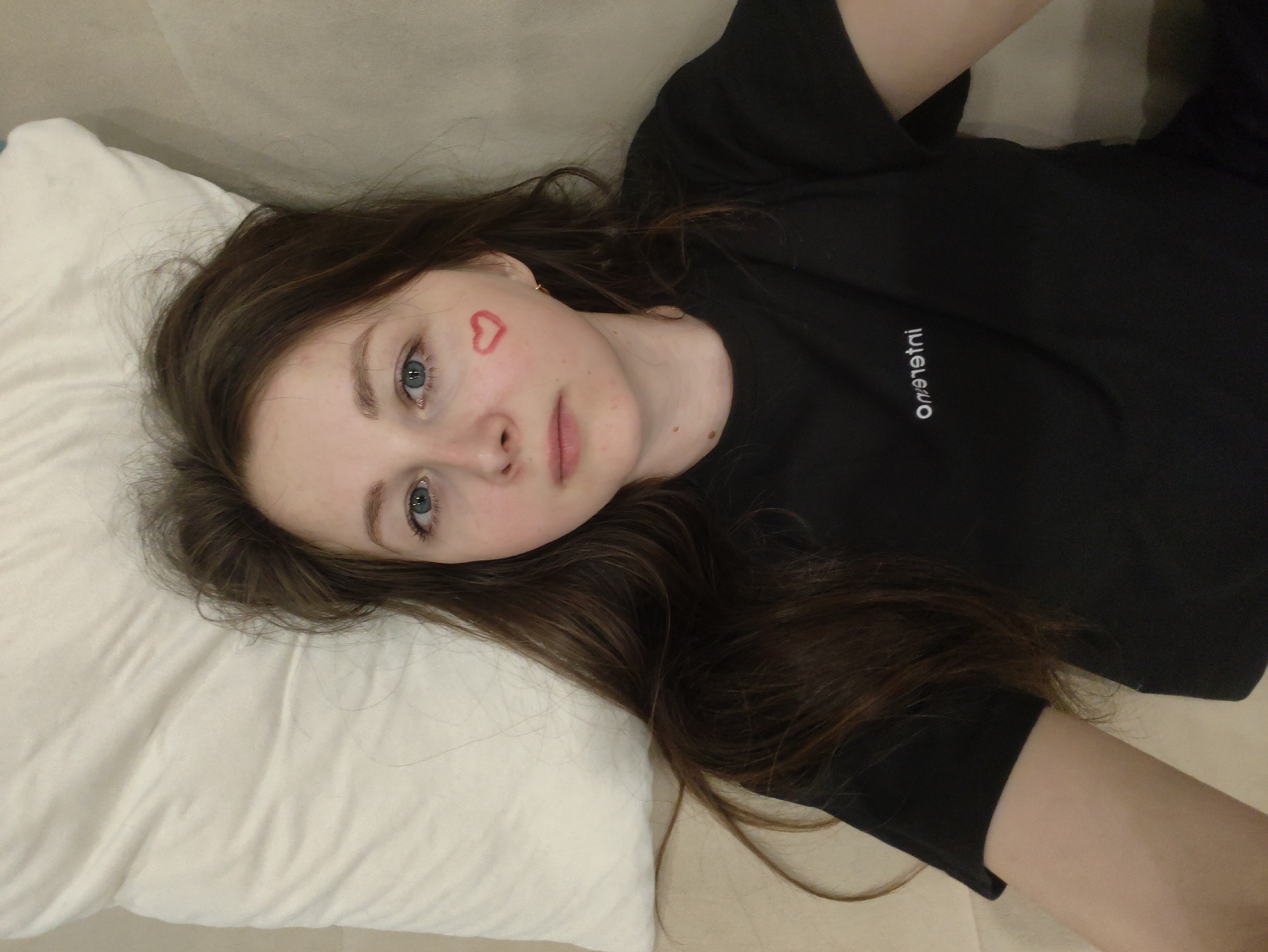 A young woman lies on a beige-colored surface, likely a couch or bed, with a white pillow supporting her head. She has fair skin, light blue eyes, and long brown hair that is spread around her on the pillow and surface. A red heart is drawn on her left cheek. She is wearing a black t-shirt with a small white logo on the upper left chest. The environment appears to be indoors and residential. The location is Unknown, Unknown.