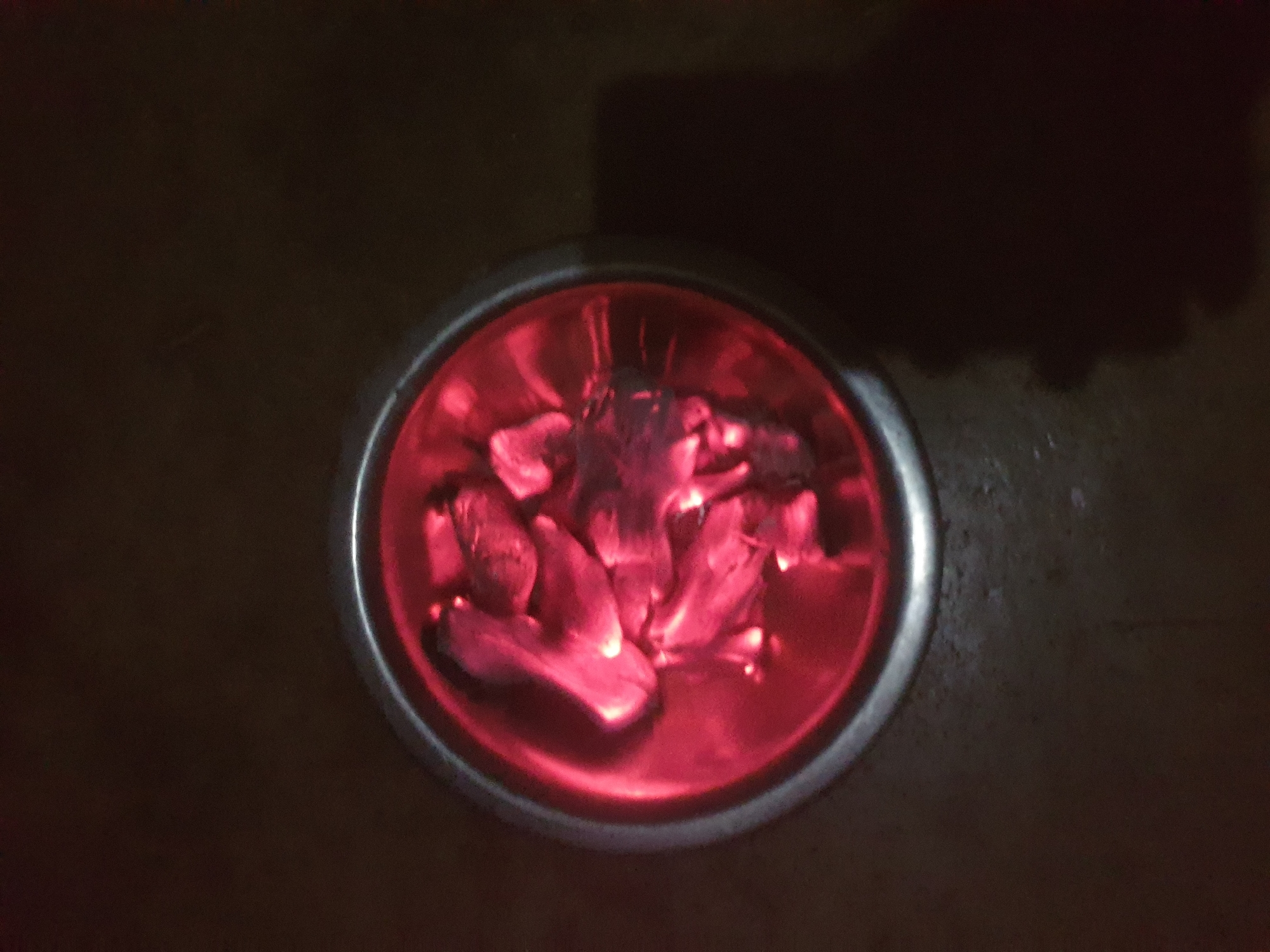 A top-down, slightly angled view captures a circular, dark metal container holding multiple irregularly shaped, incandescent red embers, indicative of burning charcoal. The container features a metallic, silver-colored rim. The intense red glow from the embers illuminates the interior of the container and casts a warm, reddish hue within its confines. The surrounding environment is predominantly dark brown-black, suggesting low ambient light conditions. A distinct, amorphous dark shadow is visible on the lower right side of the frame, contrasting with the dark background. The scene is located in the city of Unknown, Unknown.