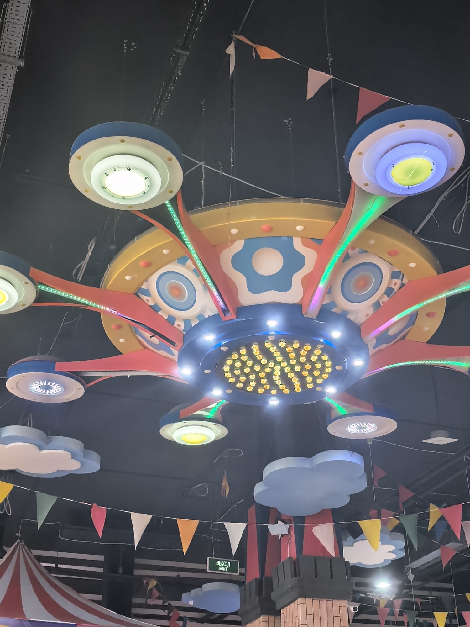 A view of a brightly colored, whimsical lighting fixture suspended from a dark ceiling. The fixture is circular with multiple "arms" radiating outwards, each capped with a round light. The arms are decorated with red, green, and other colors, and the central hub of the fixture features numerous small lights. The overall design appears to be playful and carnival-like. Below the lighting fixture, there are decorative elements including cloud-shaped pieces, banners of various colors and shapes, and a structure resembling the top of a castle wall. A tent-like structure with red and white stripes is partially visible in the lower left corner. The overall setting suggests an indoor entertainment area.