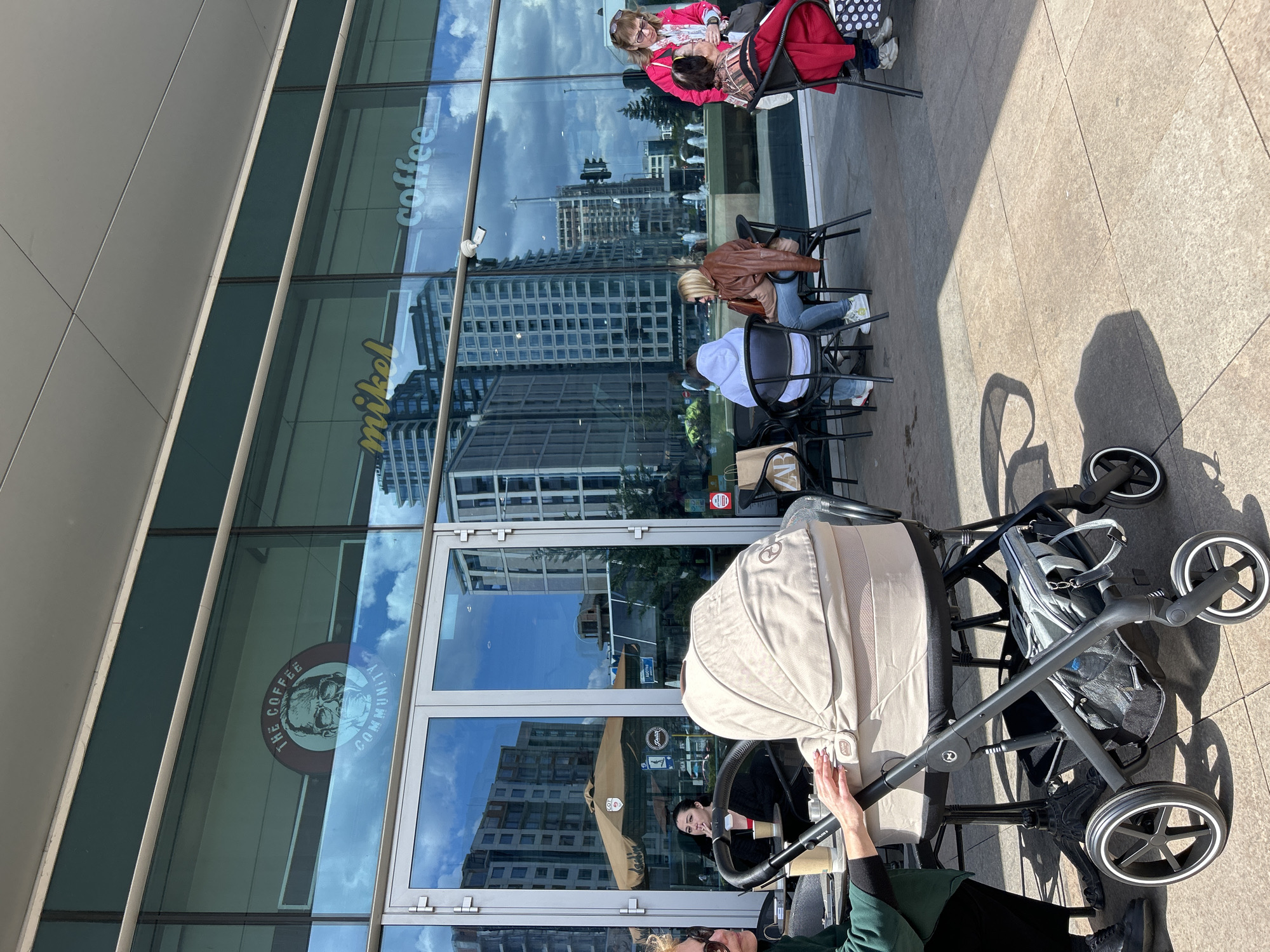 The image shows an outdoor scene in Sofia, Bulgaria, specifically near a coffee shop called "mikel coffee community" or similar. The building's glass facade reflects nearby buildings and the sky.
There are several people present, some sitting at tables outside the cafe. One group includes two women, one in a pink jacket and the other with a patterned top and dark jacket. Another group consists of three people; one wearing a brown jacket and another with a white hoodie.
In the foreground, there's a baby stroller or pram, with a light beige hood. A person wearing a green jacket appears to be interacting with it. A black diaper bag or backpack is hanging on the stroller. The ground is paved with large, light-colored tiles.