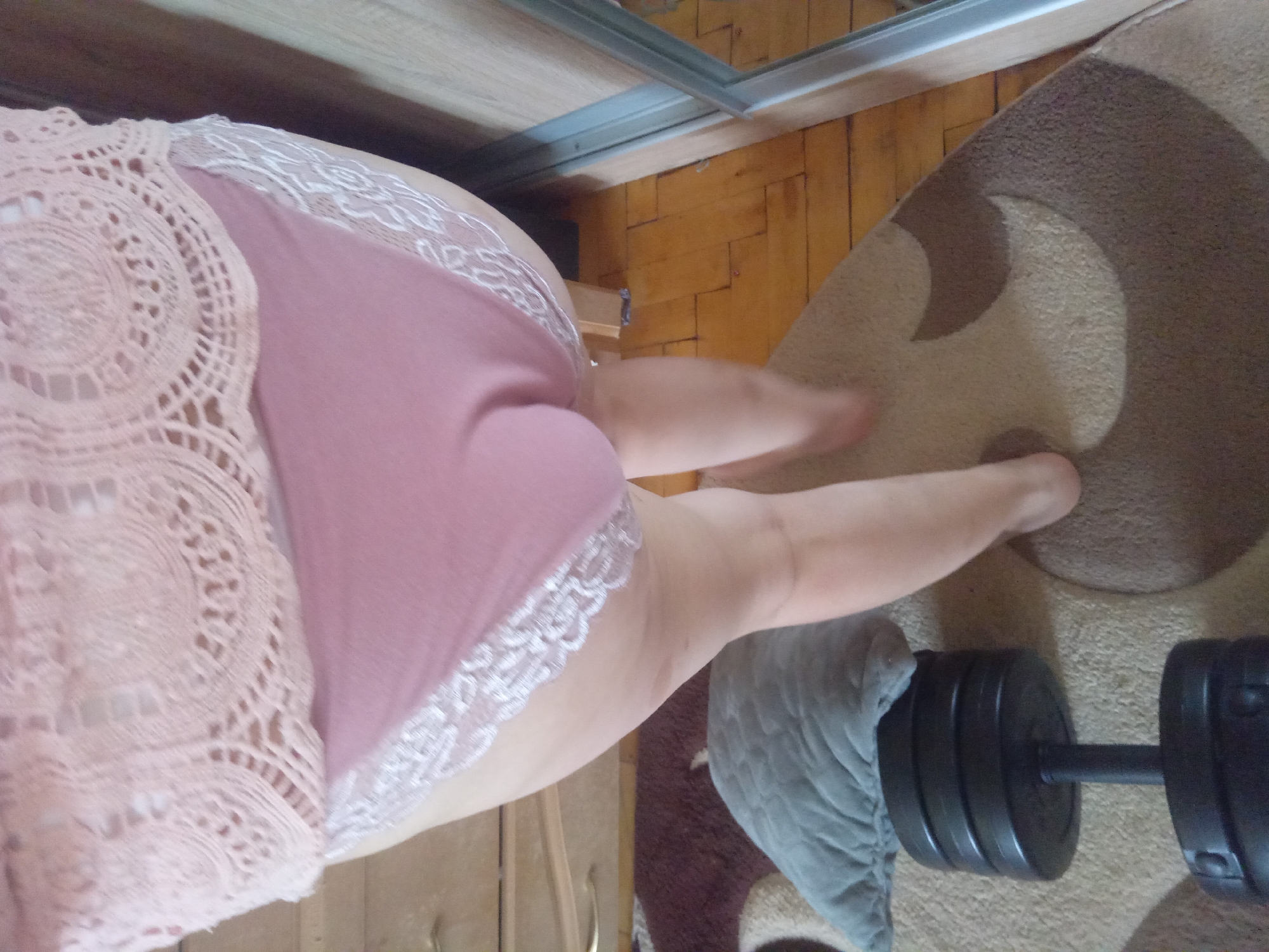 A person is standing in a room with wooden floors and a patterned carpet. The person is wearing pink lace trim underwear and a pink lace top. A dumbbell is on the carpet next to a grey pillow. A dresser is visible in the background. The image is taken from a low angle, looking up at the person. The scene is indoors and does not contain any identifiable landmarks of Terebovlia, Ukraine.