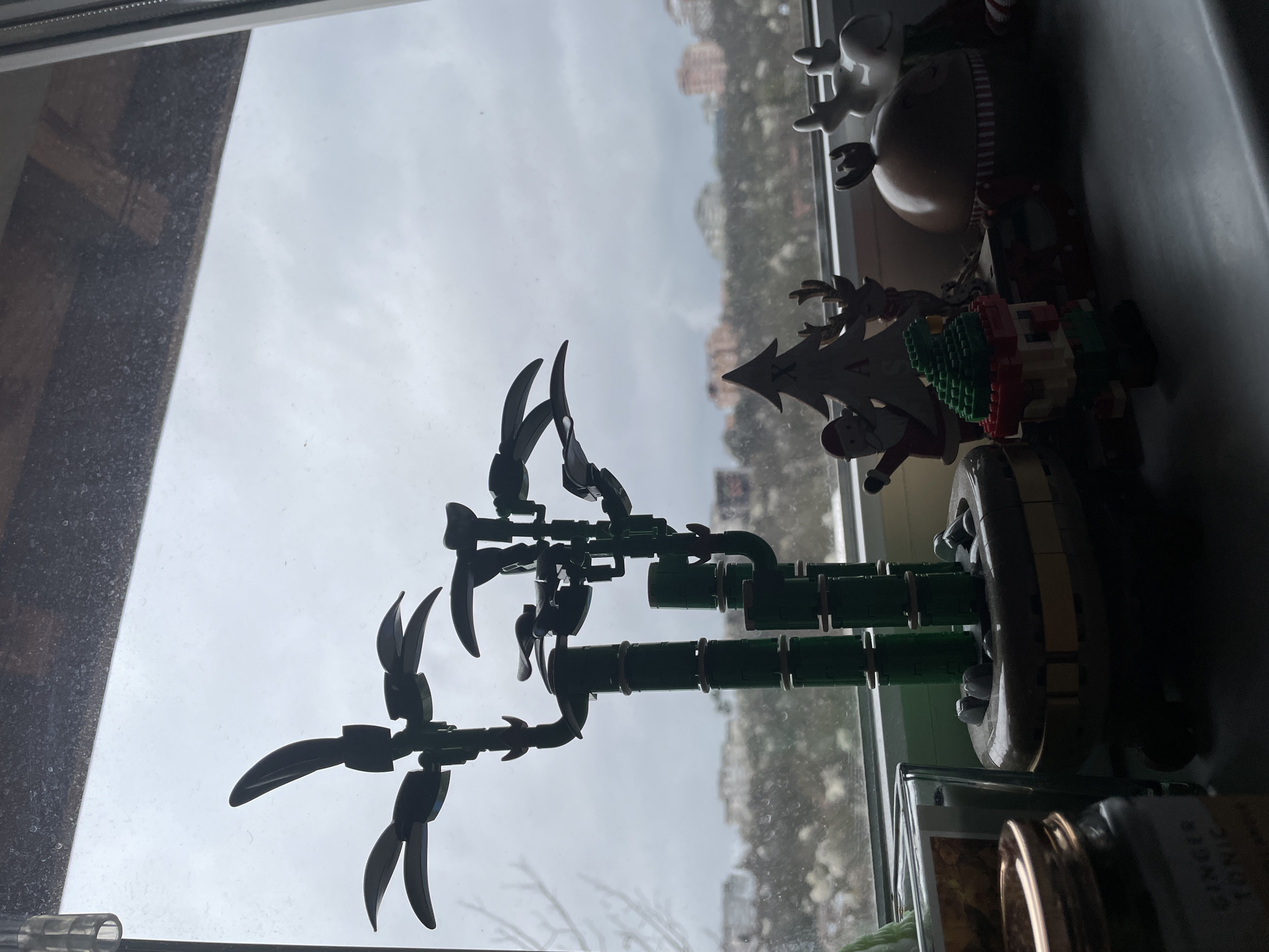 The scene is indoors, near a window, and includes a Lego tree model, other Christmas decorations, and part of a window view of the city.
Specifically, in the foreground and against the window light is a Lego model of a palm tree, constructed with dark green Lego pieces. Other Christmas ornaments include a reindeer and snowman, a Christmas tree figure with Santa, and a gold-colored container.
The window has a blurred and indistinct view of a town, which is Veliko Tarnovo, Bulgaria. The weather appears to be overcast and cloudy, but it is difficult to discern further details of buildings, people, or street activity.
