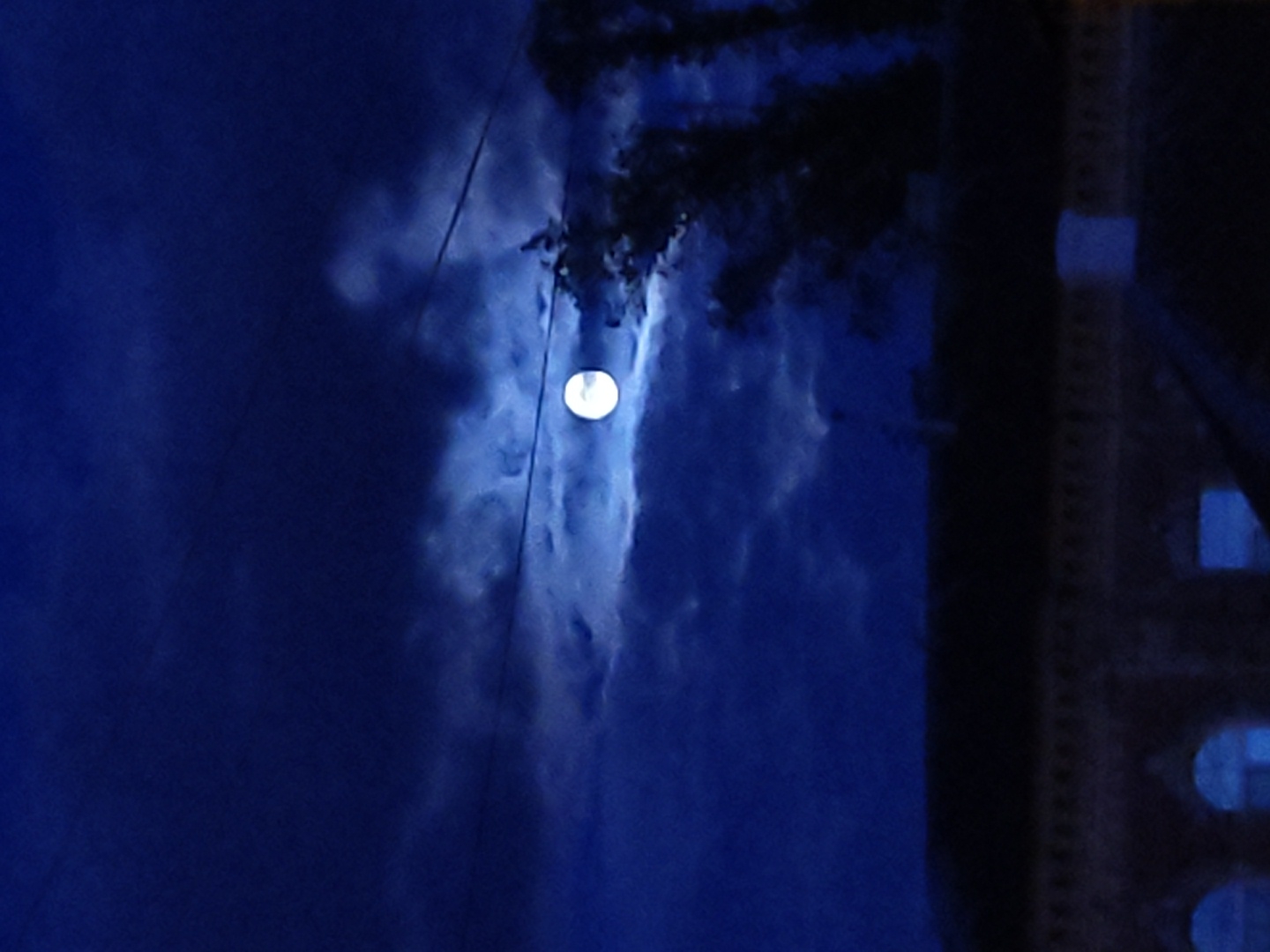 A low-angle, night-time view reveals a luminescent full moon, partially obscured by wisps of cirrus clouds. The scene is primarily cast in shades of deep blue, with the moon providing the main source of light. A power line stretches across the sky, cutting through the moon's illuminated path. Adjacent to the moon, silhouetted foliage indicates the presence of a tree. To the right, a structure appears to be the side of a building with a grid-like pattern and illuminated windows. The location is likely an urban area given the building and infrastructure.