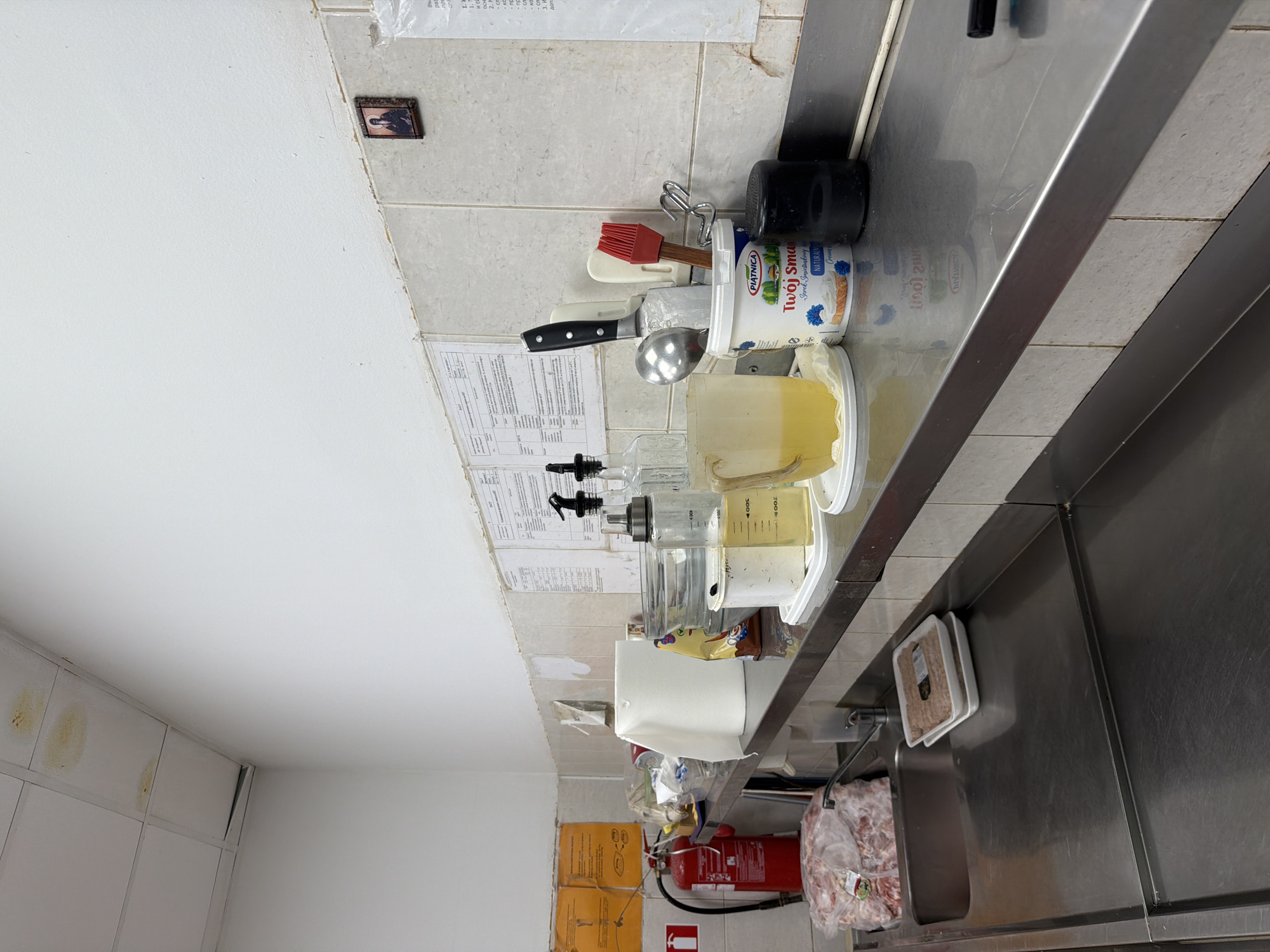 The image depicts a commercial kitchen counter with stainless steel surfaces. Various kitchen items are present, including a red fire extinguisher, large plastic bags of raw meat, a sink, packaged food items, bottles of oil, a container of yogurt, a mug with yellow liquid, and kitchen utensils such as knives and a spatula. The wall behind the counter is tiled with a light-colored tile and has some papers or notices attached. The ceiling shows some water stains and discolored tiles. The overall impression is of a functional, albeit somewhat cluttered, kitchen environment. The specific location in Plovdiv, Bulgaria, cannot be determined from the visual information alone.