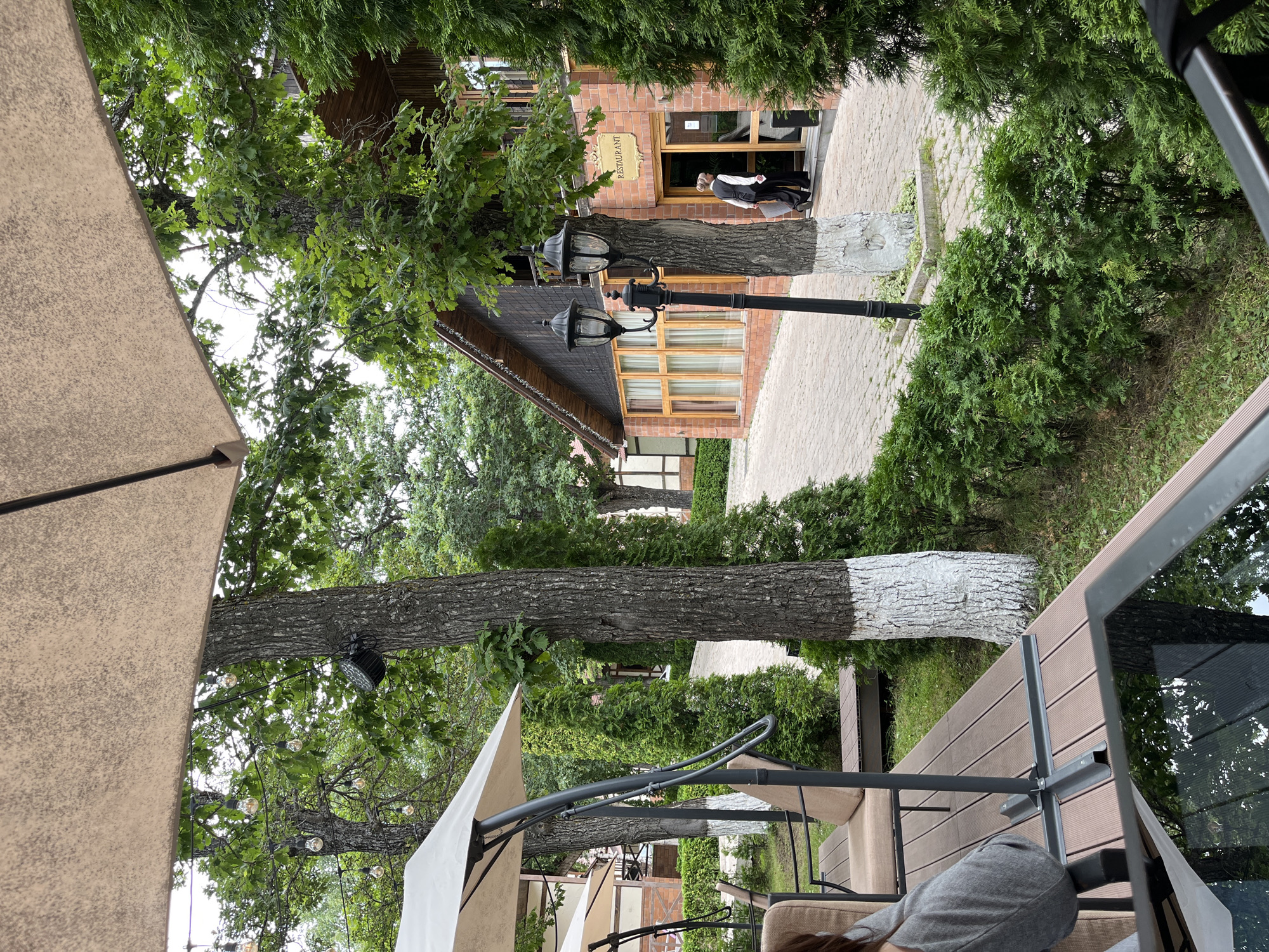 This image depicts an outdoor seating area of a restaurant in Tsaghkadzor, Armenia. In the foreground, a portion of a table with a reflective surface is visible, showing a reflection of trees. To the left, an outdoor umbrella and seating furniture, including chairs with cushions, are set up on a wooden deck. The deck is surrounded by lush green foliage, including trees and manicured hedges.
In the midground, a brick building with a dark wood trim and large windows houses the restaurant. A sign above the entrance reads "RESTAURANT". Two black lampposts stand in front of the building. A person wearing a white shirt and a dark vest is seen walking towards the entrance of the restaurant. Trees, some with trunks painted white at the base, are scattered throughout the scene, adding to the natural ambiance. The ground is paved with light-colored cobblestones. The overall environment suggests a tranquil and inviting outdoor dining space.
