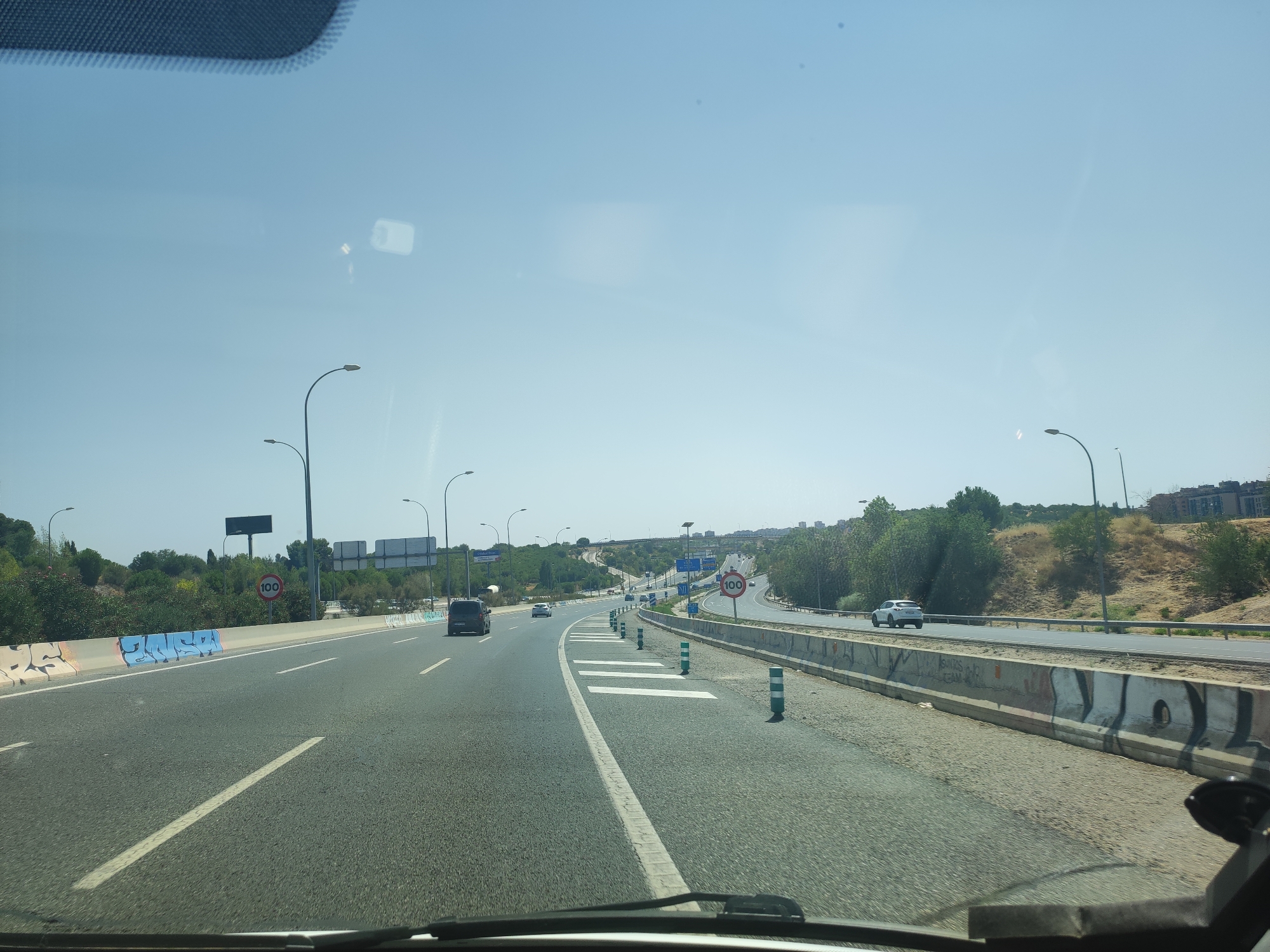The image shows a view from a vehicle driving on a multi-lane highway on a clear day. The highway has concrete barriers and multiple lanes of traffic. There are cars and vans on the road. Traffic signs are visible, including a speed limit sign indicating 100. Streetlights line the side of the highway. The surrounding environment consists of dry, grassy hills with trees and some buildings visible in the distance. The overall scene suggests a suburban or highway exit area within the city of Madrid, Spain.