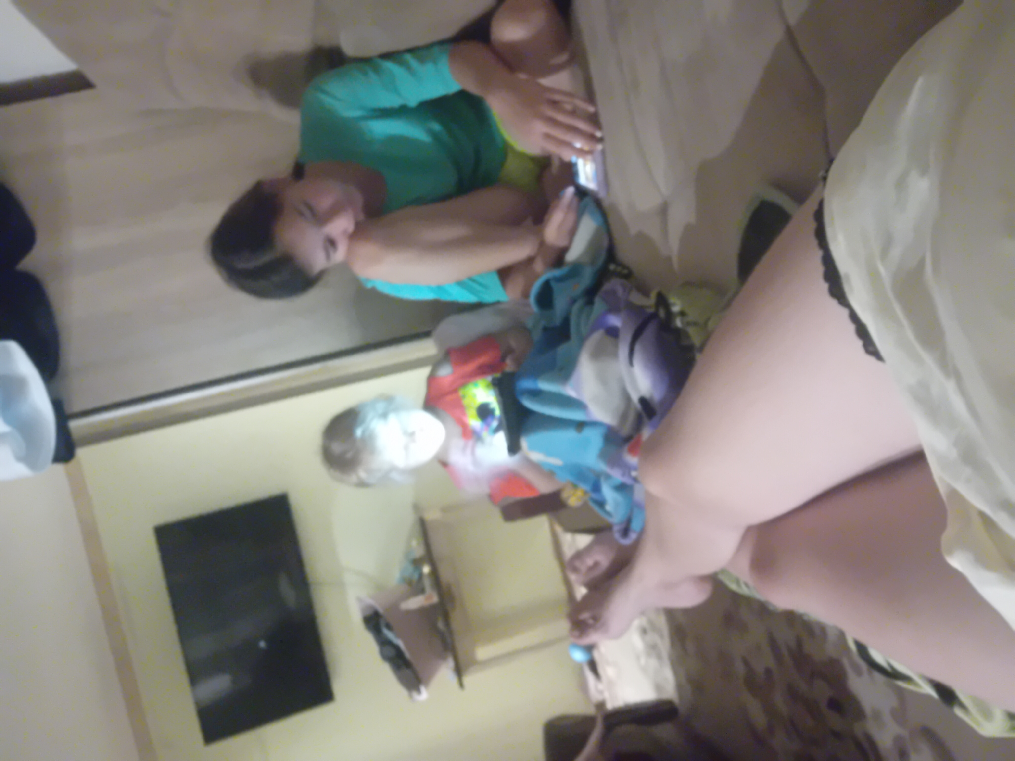 In an indoor setting, two young females are seated on a couch, engrossed in electronic devices. The female on the right, wearing a teal shirt and yellow shorts, is looking down at a tablet or phone. The younger female to her left, wearing a red shirt, is also looking at a device, illuminated by its screen. A television is mounted on the wall in the background, and a table with various items is to the left of the younger female. The overall scene depicts a casual, relaxed atmosphere within a home. The image is undated and unlocated, and therefore a specific location in Chuhuiv, Ukraine cannot be determined.