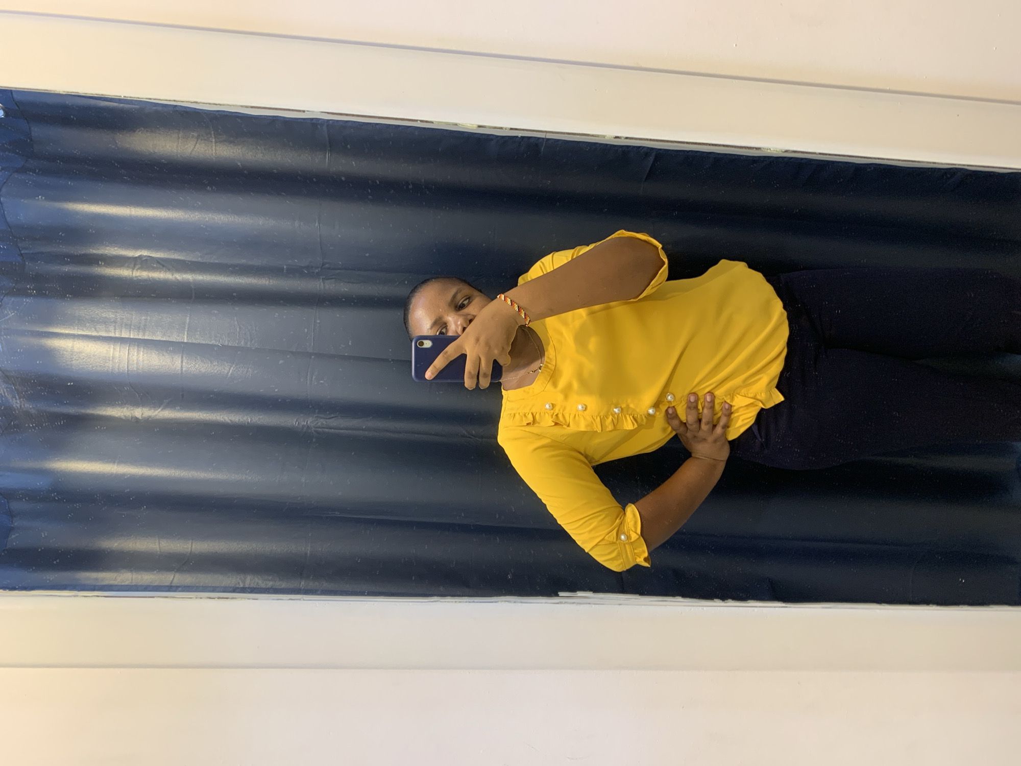 A person is taking a picture with a cell phone. The person is wearing a yellow shirt and dark blue pants. They are standing in front of a dark blue curtain. The location appears to be an interior space. There is no information about the city of Dar es-Salaam, Tanzania.