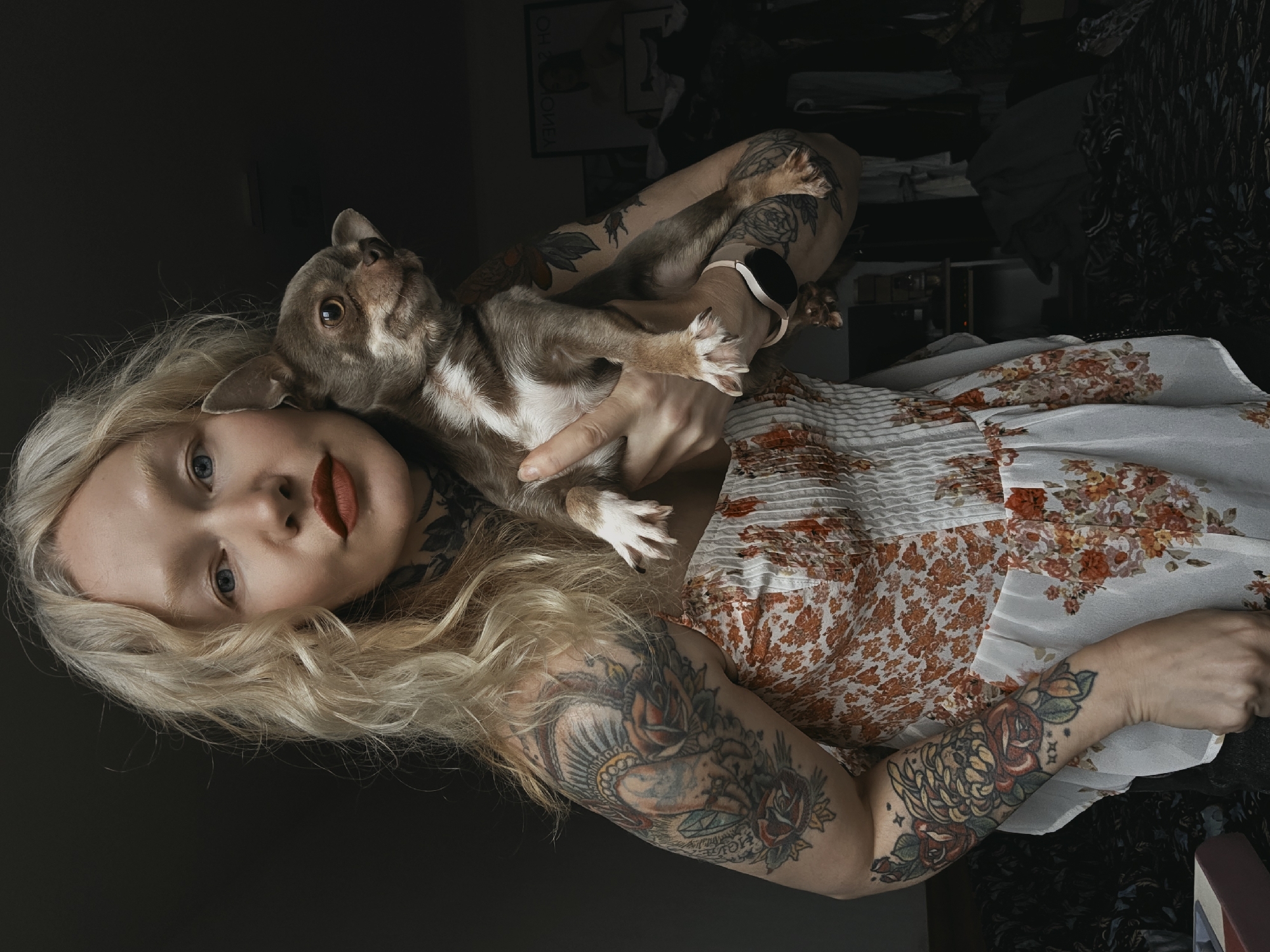 A fair-skinned woman with blonde hair is holding a brown and white dog. She is wearing a white dress with an orange floral pattern. Both of her arms are covered in tattoos. She is lying on a bed in a room that appears to be dimly lit, with items in the background, likely clothes. No clear indications suggest the location is in Saint Petersburg, Russia.