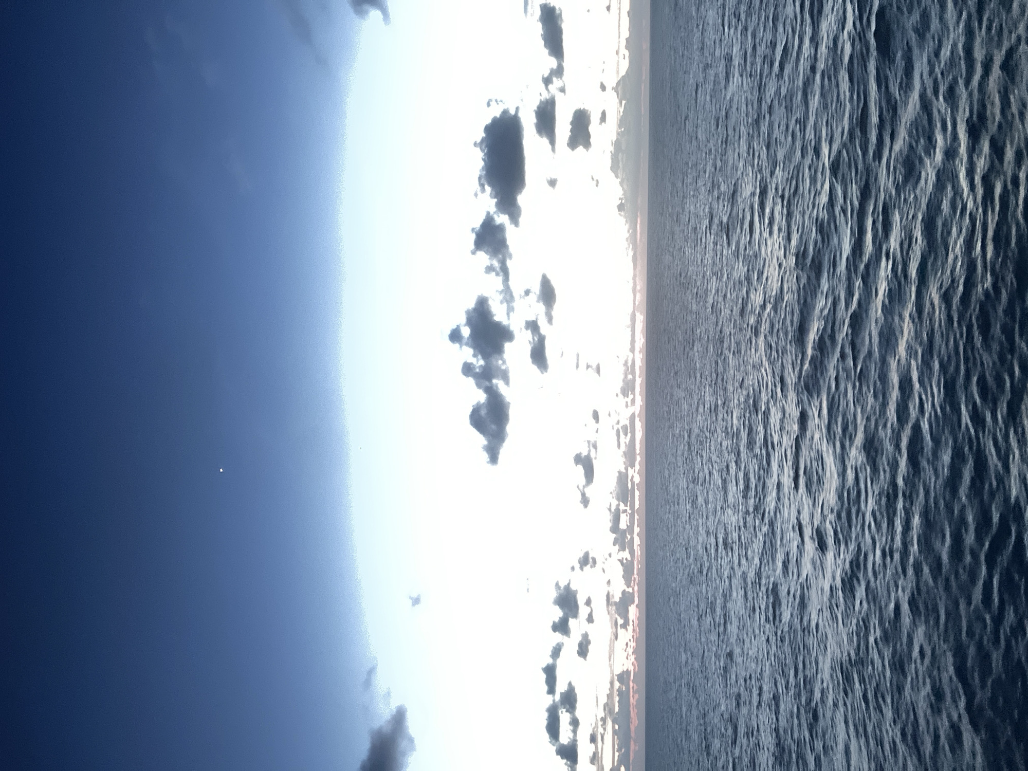 A wide-angle, eye-level shot shows a vast expanse of deep blue ocean under a dusky sky. The water's surface is textured with ripples, reflecting the muted light. In the distance, a thin band of pale pink and orange hues peeks through a line of scattered, dark gray clouds hugging the horizon. Above the clouds, the sky transitions from a bright white to a deep, clear blue, with a single, faint white dot, possibly a star or planet, visible in the upper left quadrant. The scene is tranquil, with no discernable human presence or man-made objects.