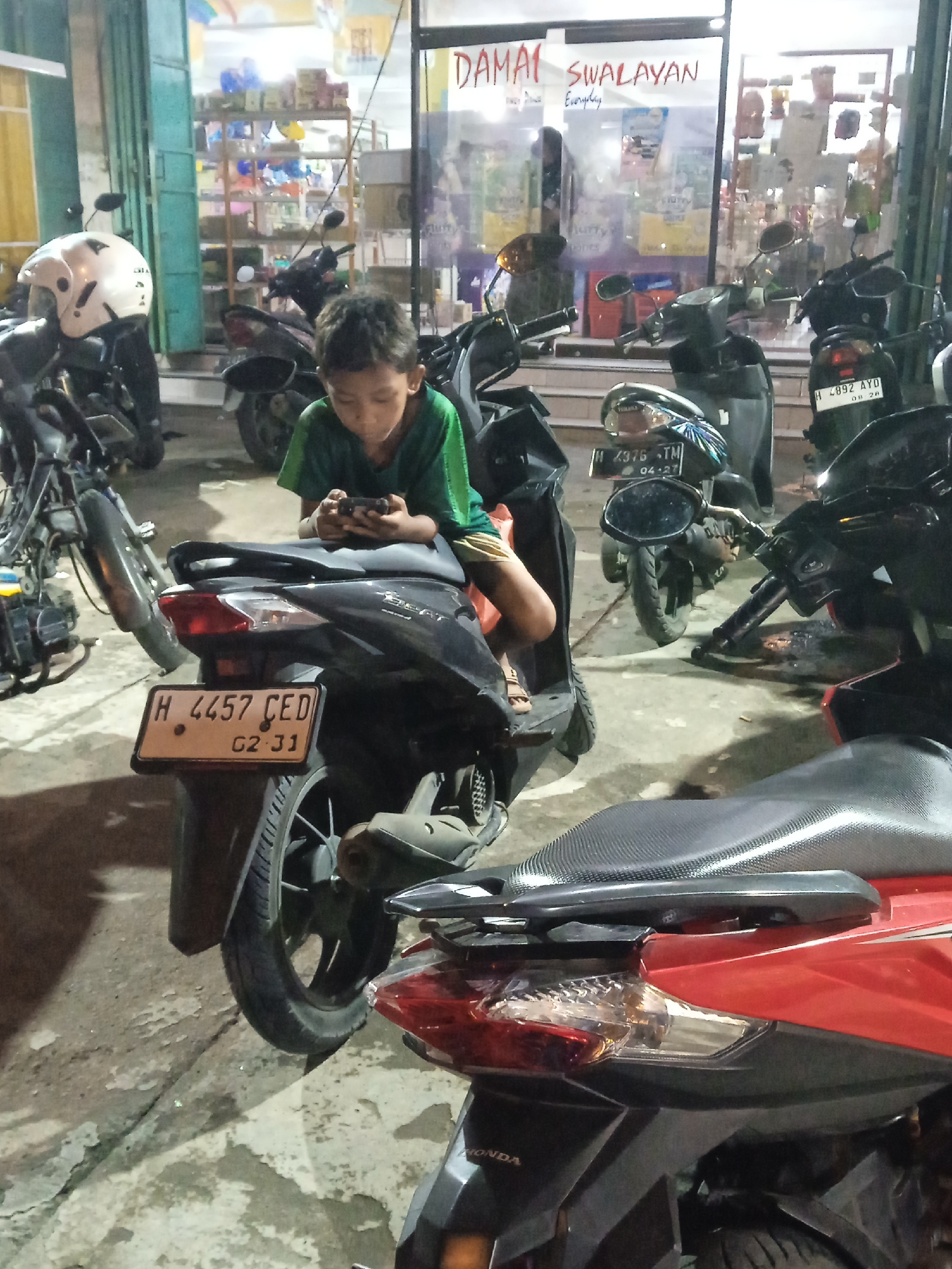A photograph taken in Kebonsari, Indonesia, shows a young boy in a green shirt sitting on a black motorbike and looking at a smartphone. The motorbike has a license plate that reads "H 4457 CED G2-31". The motorbike is parked in front of a building with a sign that reads "DAMAI SWALAYAN". Other motorbikes are parked nearby. The license plate of another motorbike visible in the shot reads "H 4376 TM 04-27". Another license plate reads "H 4892 AYD 08 28". A red Honda motorbike is also partially visible in the foreground. The ground appears to be concrete. The store behind the motorbikes has items displayed in shelves and on a rack, suggesting a convenience store.