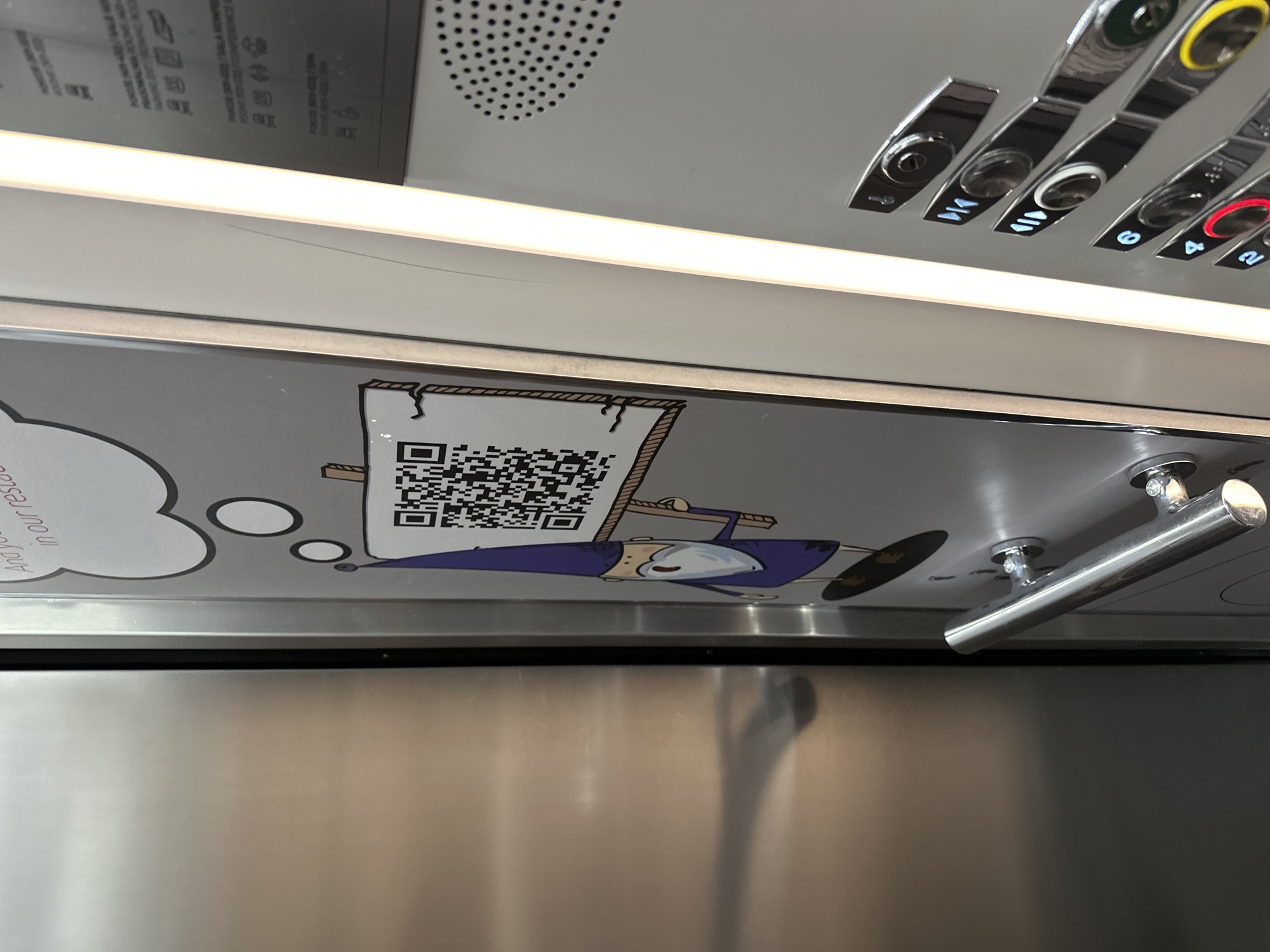 The image is taken inside an elevator. It shows a close-up of a gray panel above the elevator door, containing a cartoon illustration of a character holding a banner with a QR code on it. Above the panel are elevator buttons for floors 8, 6, 4, and 2, as well as buttons with arrows for up and down. To the right of the cartoon are the elevator's handrails. Based on the distinctive design visible on the panel, this is likely an elevator located within the Novotel Wrocław Centrum hotel in Wrocław, Poland.