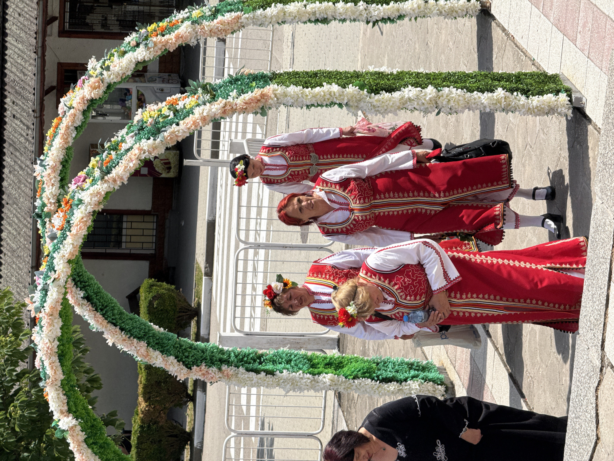 In Pordim, Bulgaria, four women dressed in traditional Bulgarian folk costumes are walking through a floral archway. The costumes are predominantly red and white, with intricate embroidery and gold trim. Two of the women are wearing floral headpieces. The archway is constructed with green foliage and adorned with numerous artificial flowers in shades of white, pink, and orange. Behind the archway, a street with a pedestrian crossing is visible, along with buildings and some greenery. The scene appears to be outdoors, under a bright sky, suggesting a public event or celebration.