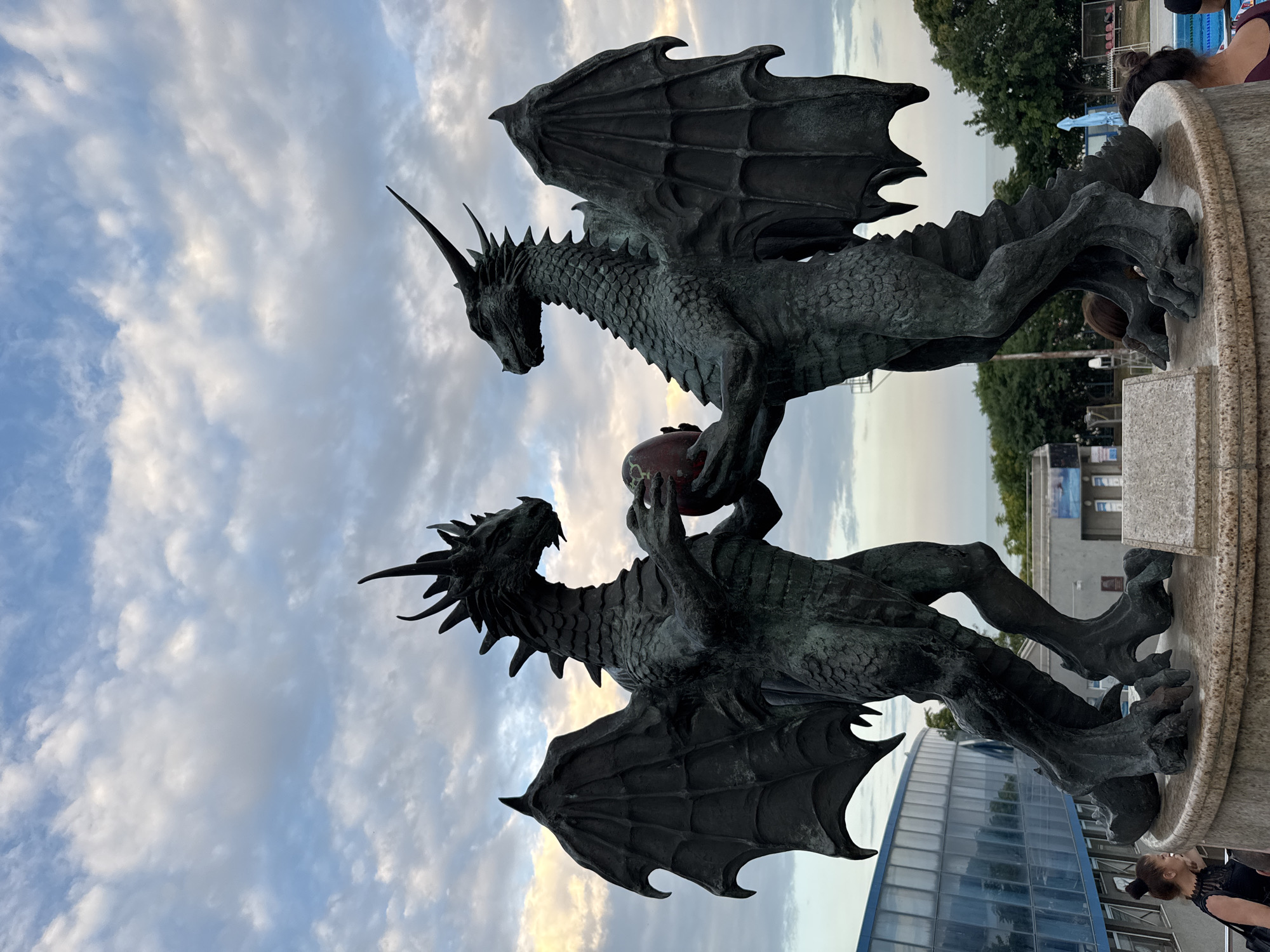 This media file features a low-angle shot of two intricately detailed bronze dragon sculptures positioned on a pedestal. The dragons are depicted in a dynamic pose, with one dragon appearing to offer a reddish-brown orb to the other. The sculpture is set against a sky filled with dramatic clouds, suggesting either dawn or dusk. In the background, glimpses of a modern building with a glass facade and trees can be seen, indicating an outdoor, likely urban, setting. A few people are partially visible near the edge of the frame, suggesting this is a public space. The overall impression is one of mythical grandeur and artistic craftsmanship. The scene is likely in Varna, Bulgaria, given the location context.