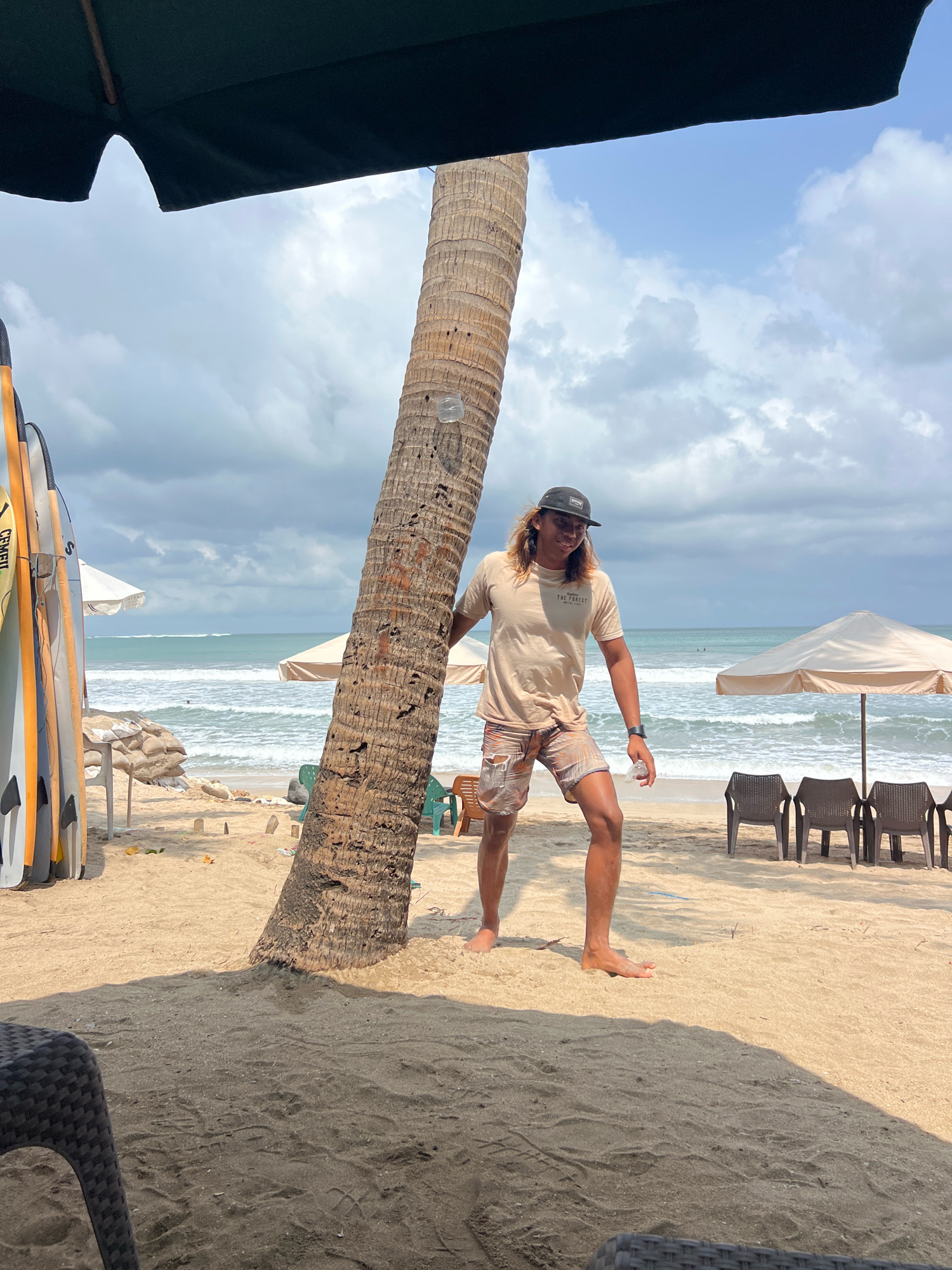 In Kuta, Indonesia, a man with long, blonde hair, wearing a tan t-shirt with a graphic print and patterned shorts, stands on a sandy beach leaning against a palm tree. He is wearing a black baseball cap and sunglasses. To his left, a row of surfboards stands upright. In the background, the ocean is visible with waves breaking. Beach umbrellas and chairs are set up on the sand. The sky is partly cloudy.