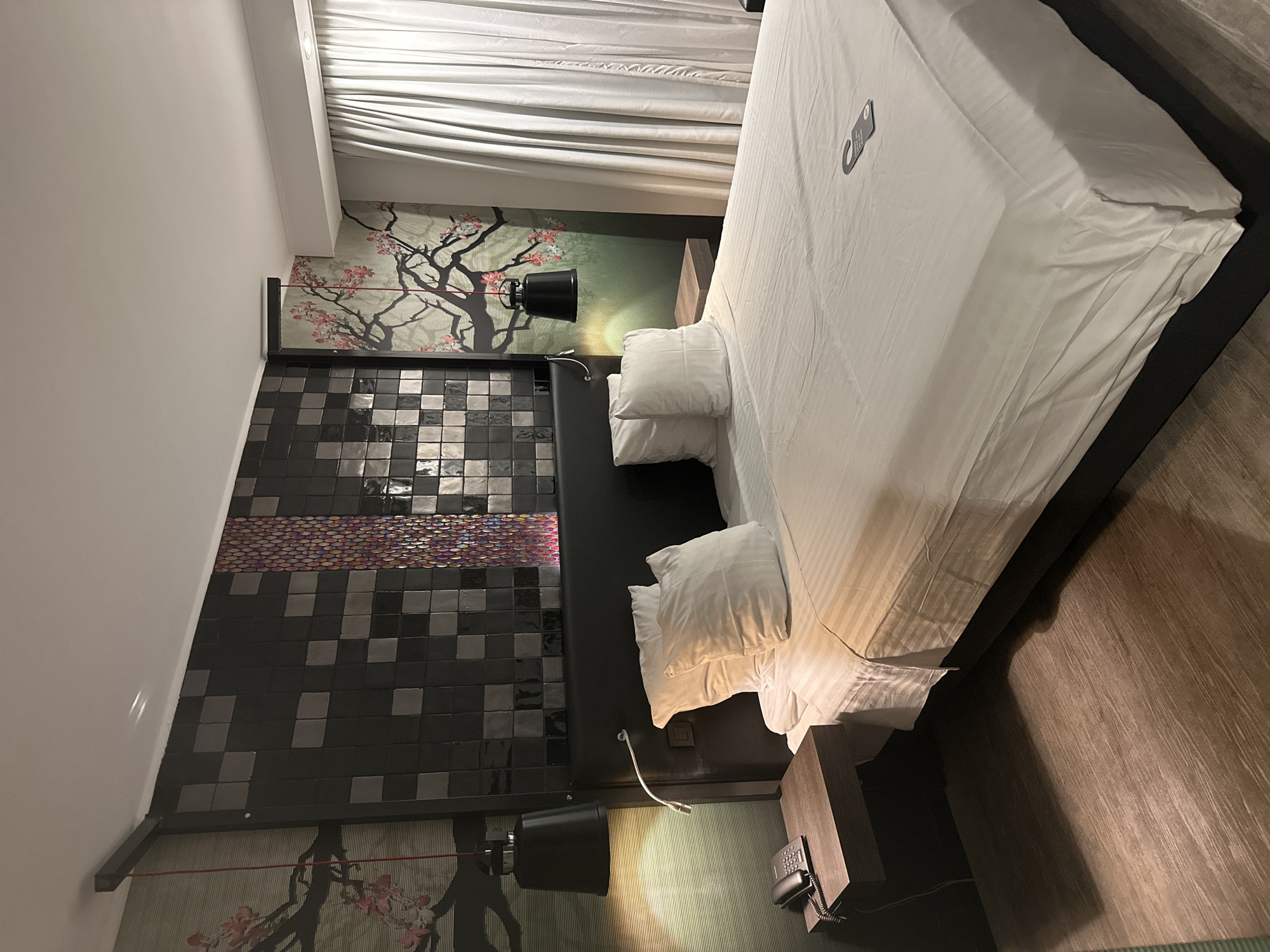 A bedroom interior is depicted, likely in a hotel in Genk, Belgium. The room features a bed with white linens and pillows, positioned against a headboard tiled with black and gray squares with a section of colorful round tiles. A wallpaper mural showing a tree branch design is visible behind the headboard, and there are black lamps mounted on either side of the bed. A bedside table is located on one side of the bed, holding a telephone. The floor appears to be wood-like. The room is illuminated, with daylight coming in through the curtains.