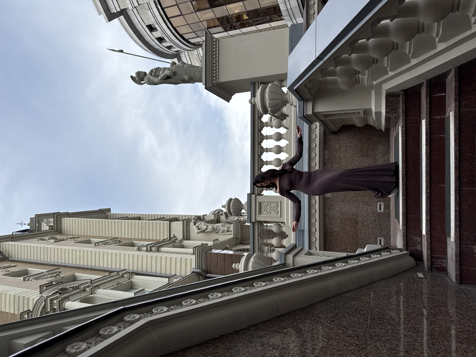 A woman in a long, dark purple dress stands on a wide, red-tiled staircase with ornate white balustrades. She is positioned on a lower landing, her arms extended to her sides, palms facing upward. The staircase is flanked by buildings with classical architectural elements. To the left, a building features a facade with alternating light and dark stripes, ornate window frames, and decorative sculptures. To the right, a modern building with large glass windows is visible, and a statue of a warrior holding a spear stands atop a pedestal. The sky above is overcast and cloudy. The overall scene suggests an urban setting, likely a public square or the entrance to a grand building. The architectural style and the statue hint at a location with historical significance. The woman's pose is elegant and dramatic, drawing attention to her and the surrounding architecture. There is no immediate indication of the city of Kazan, Russia, within the visual elements of the image.