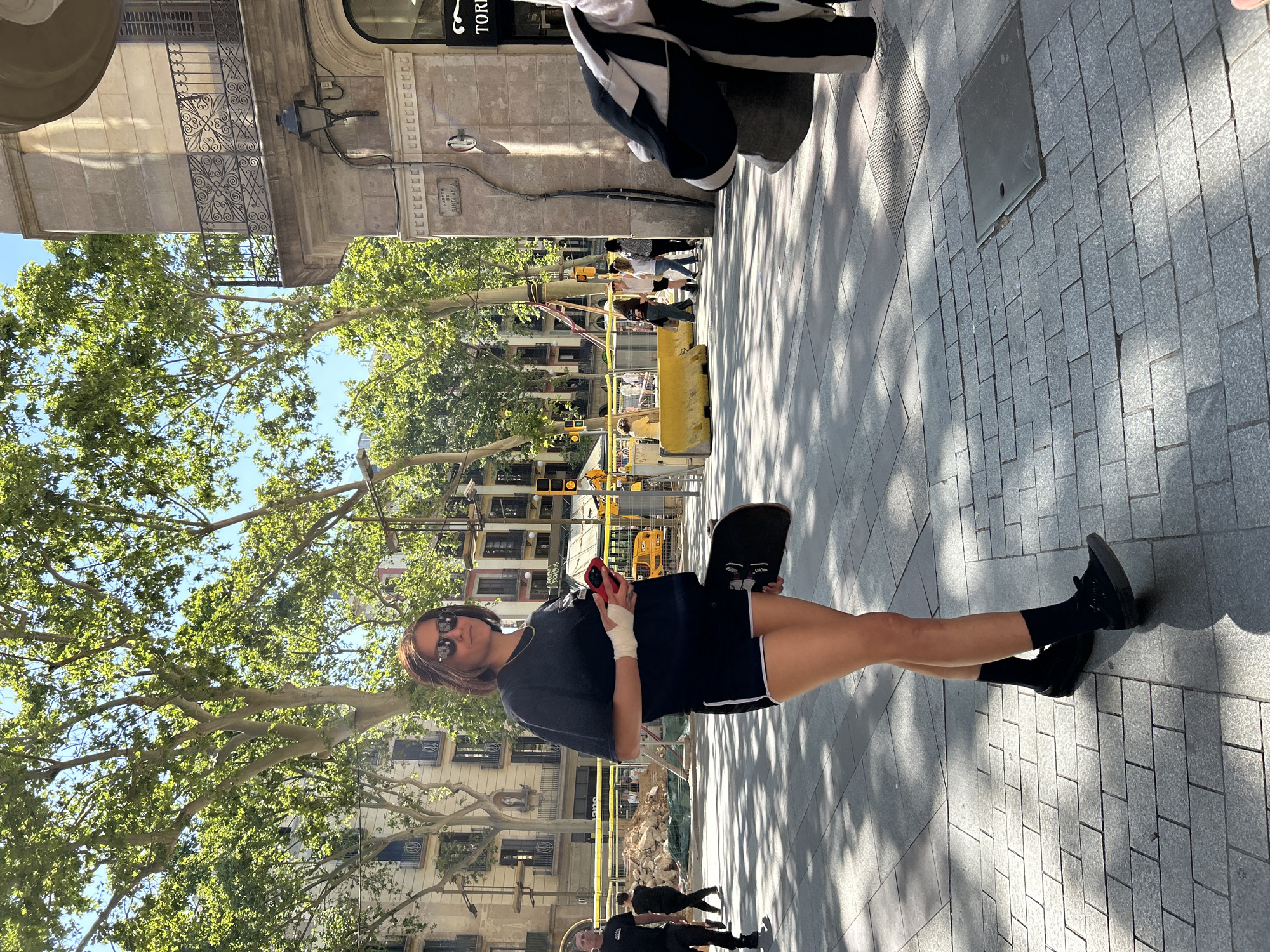 A woman stands on a gray-paved sidewalk in Barcelona, Spain, holding a skateboard in her left hand and a cell phone in her right. Her left hand is bandaged. She wears a dark t-shirt, dark shorts with white trim, black socks, and black sneakers. She has sunglasses on. In the background are buildings with multiple windows and trees. Yellow construction equipment and barricades are visible further in the background. Other pedestrians can be seen in the distance. A street sign with the text "Carrer de Santa Anna" is affixed to the building on the right side of the image.