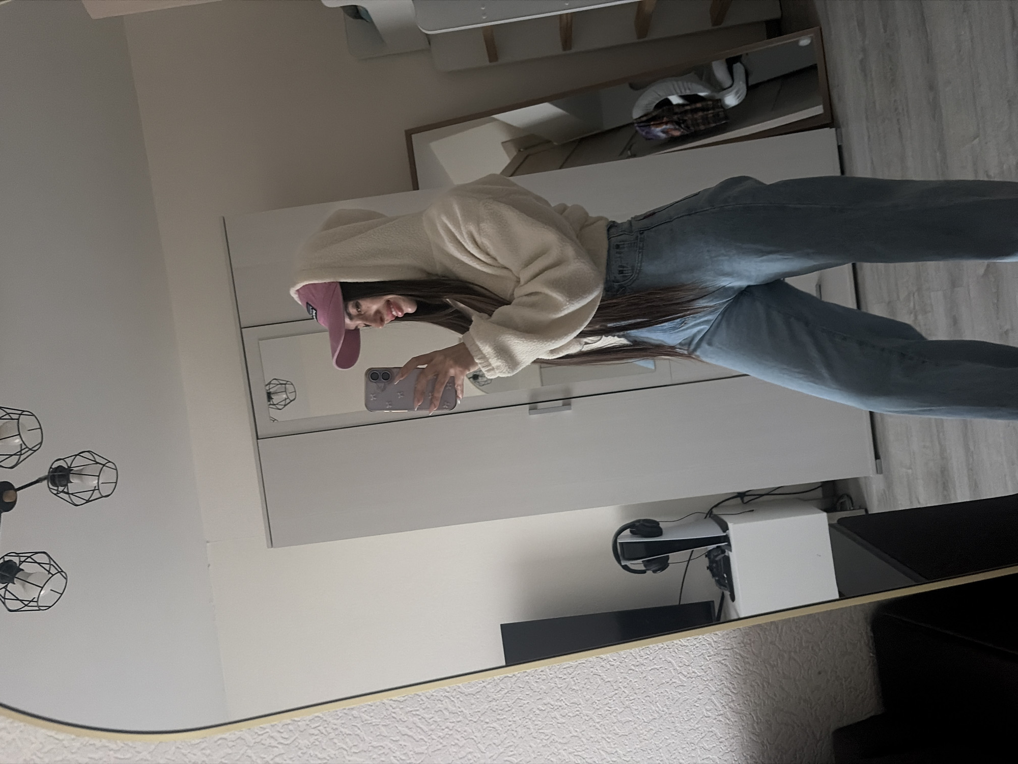 The image shows a person reflected in a mirror. The person is wearing a pink baseball cap, a light-colored hooded jacket, and blue jeans. They are holding a smartphone, presumably taking the photo. In the background, there is a white cabinet or wardrobe, a television set, and a gaming console with headphones. A modern light fixture is also visible in the background. The location is not discernible from the image.