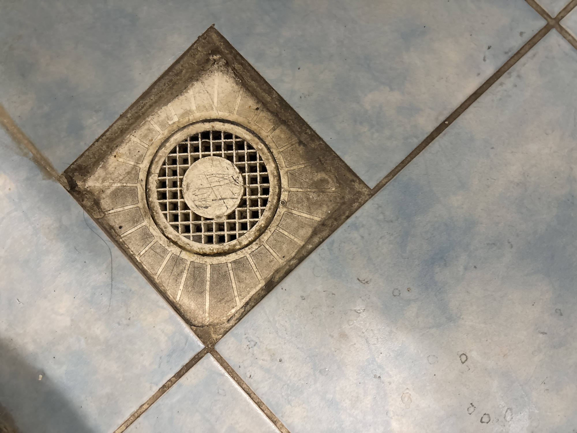 The image shows a close-up, overhead view of a floor drain set into light blue square tiles. The drain is square with a circular grate and is surrounded by a dirty, gray concrete fixture. The tiles are laid out diagonally, and grout lines are visible between them. Some minor dirt and scuff marks are present on the tiles around the drain. The lighting is overhead, casting subtle shadows. No people or actions are depicted. The location is described as Unknown, Unknown, which implies no specific city or place is identified.