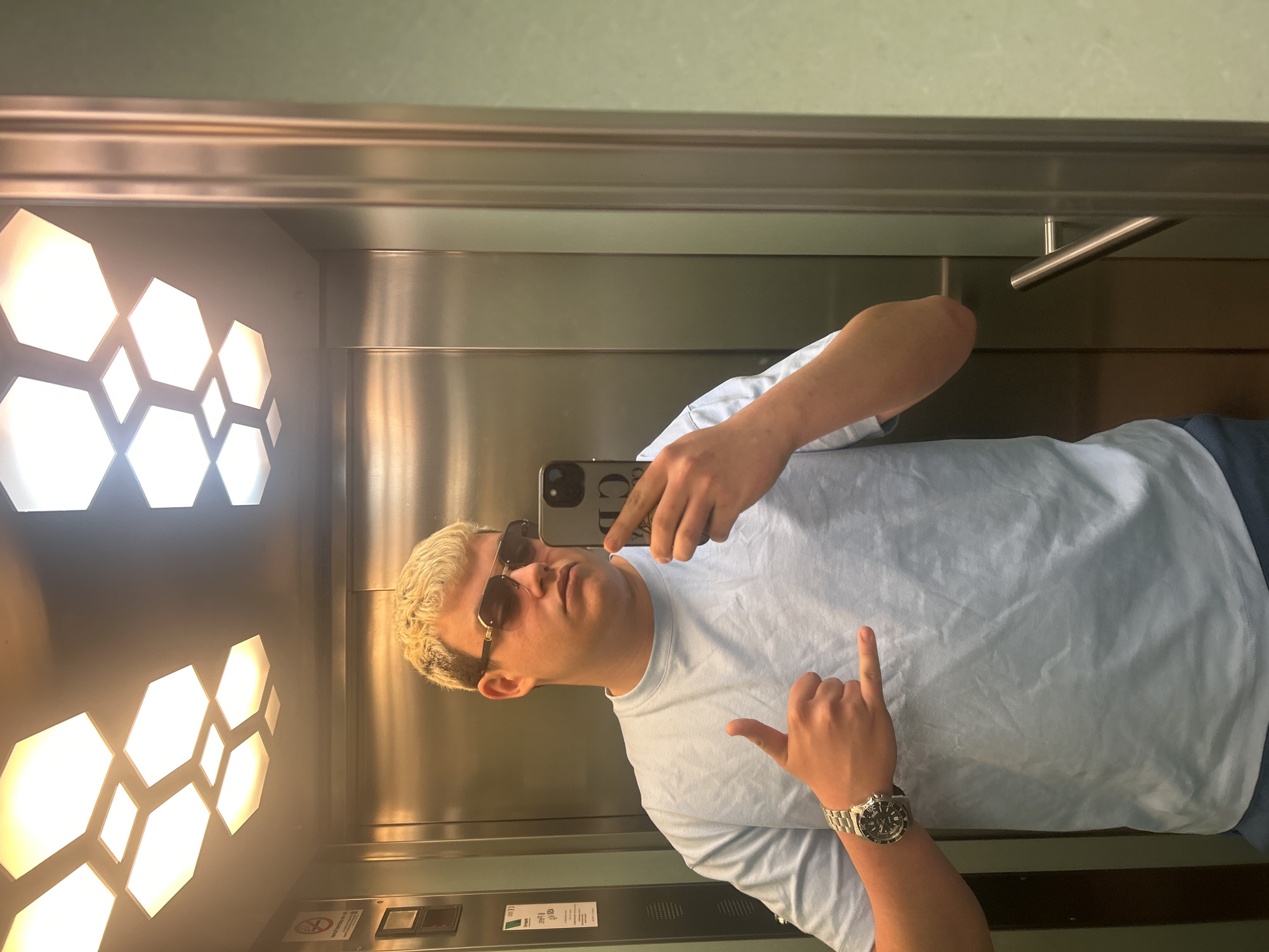 A man stands in an elevator, taking a selfie with a smartphone. He is wearing a light blue t-shirt, sunglasses, and a watch. He has short blonde hair and is making a hand gesture. The elevator has hexagon-shaped lights on the ceiling. The location is possibly in Lleida, Spain, but there is no specific indicator of location within the image. The elevator interior is metallic with horizontal grooves and a stainless-steel handle. A control panel is visible on the wall near the floor.