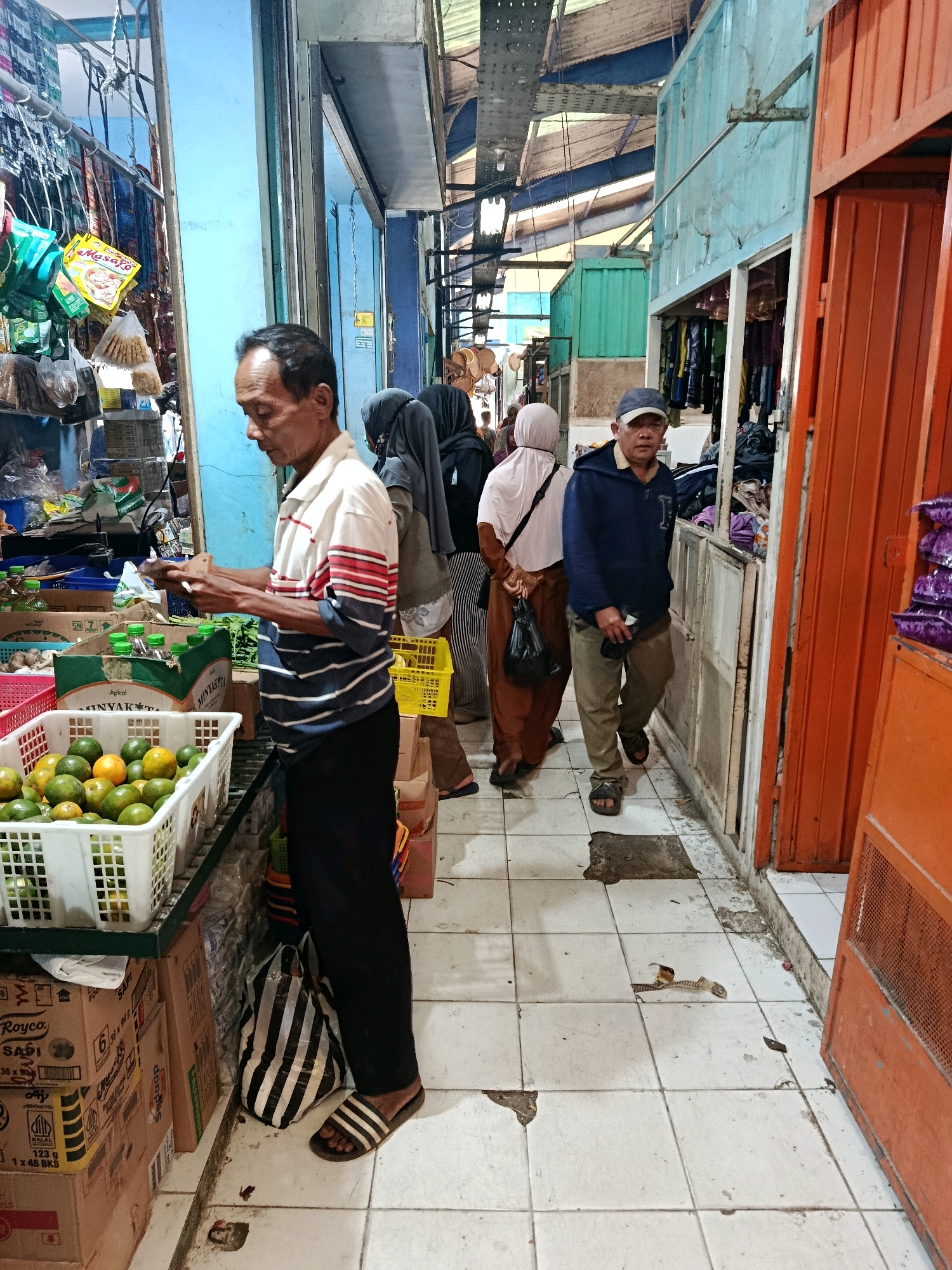 A medium shot captures a bustling market scene in Bululawang, Indonesia. In the foreground, an Indonesian man, middle-aged, with dark hair and a tanned complexion, wears a striped polo shirt and black pants. He is holding a mobile phone in his right hand and appears to be examining it. To his left, a white plastic crate overflows with fresh oranges, and behind it, boxes are stacked. The man is facing the viewer's left, slightly angled away.
Behind the man, a woman wearing a hijab and a light-colored headscarf walks away from the camera. She is carrying a black plastic bag. Further back, another person in a hijab is visible, and a man wearing a blue hoodie and a grey baseball cap walks towards the viewer's left, looking straight ahead.
The environment appears to be an indoor or covered market with stalls lining a narrow walkway. The stalls are constructed from various materials, including blue painted metal panels, and corrugated metal roofing is visible overhead. The floor is tiled with off-white squares, some of which are chipped and worn. Various goods are displayed on shelves and in crates, including packaged items, produce, and clothing. The overall impression is one of a lively and functional marketplace.