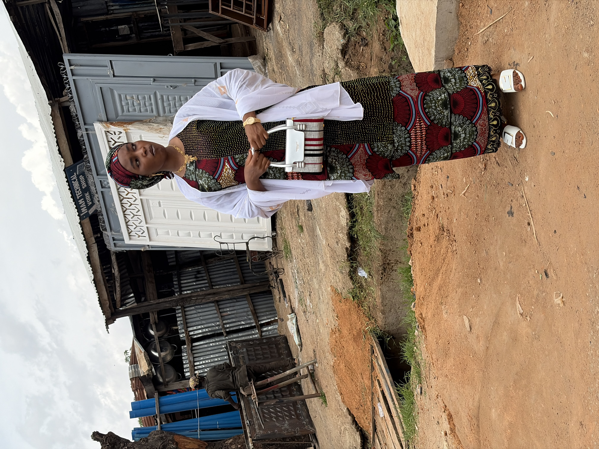 A woman is standing on a dirt road in front of a workshop. The woman is wearing a colorful dress, a headscarf, and a white shawl. She is holding a small handbag. The workshop appears to be a metal fabrication or repair shop, with corrugated metal siding and a tin roof. Various metal items, including what appear to be doors or gates, are visible in and around the workshop. In the background, there are blue pipes, large metal pots, and construction materials. The ground is predominantly dirt and sand, with some scattered debris and patches of grass. A person wearing a dark shirt and a cap is working at a metal table in the background. The sky is overcast. The location is identified as New Stadium, Nigeria.