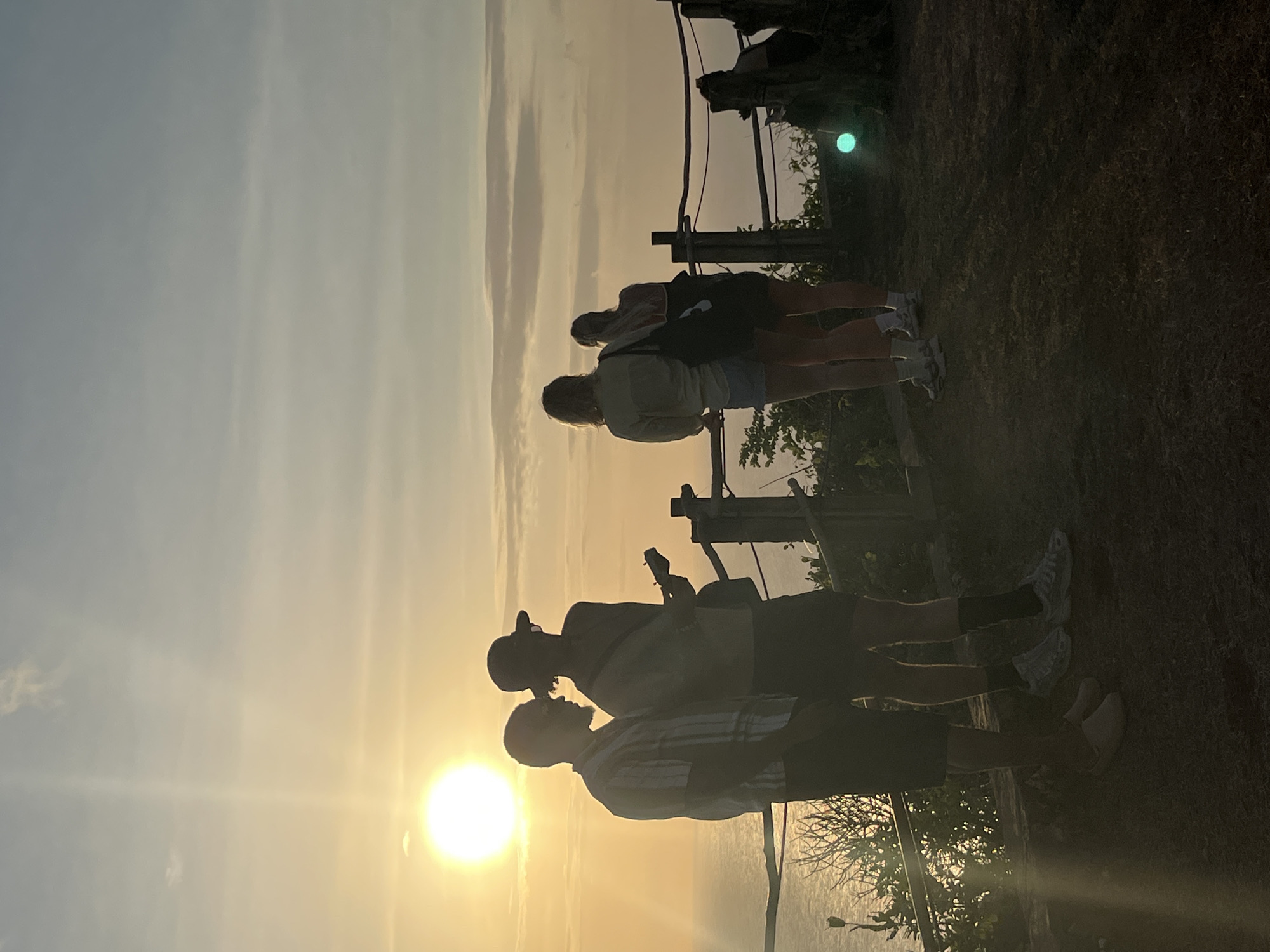 The image captures a group of people silhouetted against a golden sunset. In the foreground, two men are visible, one wearing a striped shirt and the other a cap. One of the men appears to be holding a guitar, suggesting a musical activity. Behind them, a fence runs horizontally across the frame. Further back, two women stand facing away from the camera, also silhouetted. The setting appears to be an elevated viewpoint, possibly a cliff or a hill, with vegetation visible in the background and a hazy landscape stretching towards the horizon. The sun is low in the sky, casting a warm glow and creating lens flare. The overall mood is serene and contemplative, with the soft light and the presence of people enjoying the view. The time of day is clearly sunset. There is no visible text in the image. The provided location context places this scene in Kuta Selatan, Indonesia, suggesting a tropical coastal environment.