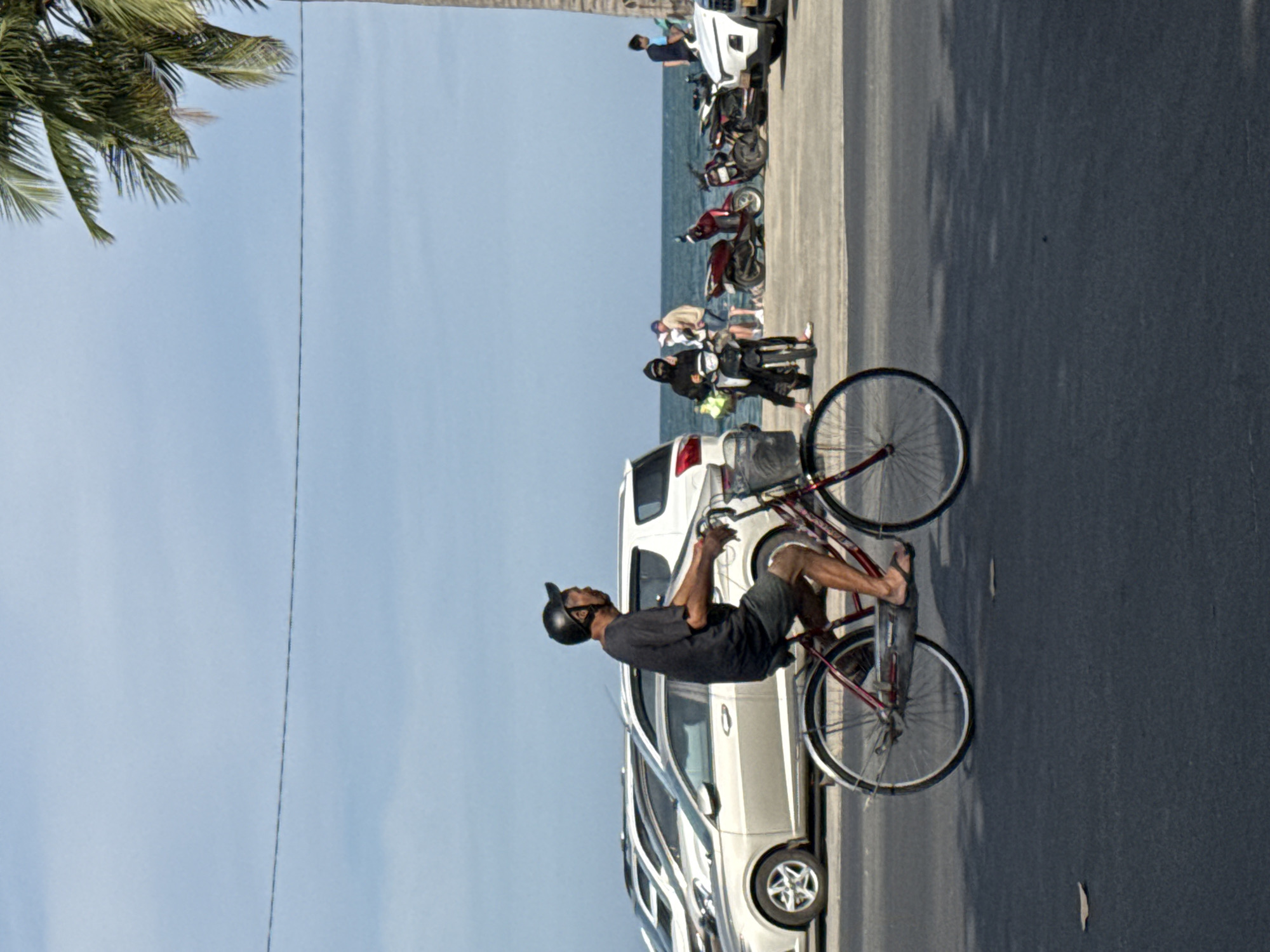 A man wearing a dark t-shirt, dark shorts, and a black helmet rides a red bicycle on a paved road. The bicycle has a basket on the front and a rack on the back. He is wearing flip-flops. In the background, there are several cars parked on the side of the road, and further back, the ocean is visible with a few motorcycles parked near the water. A palm tree is on the right side of the frame, and a power line stretches across the sky. The sky is clear and blue. The scene appears to be in a coastal city in Vietnam.