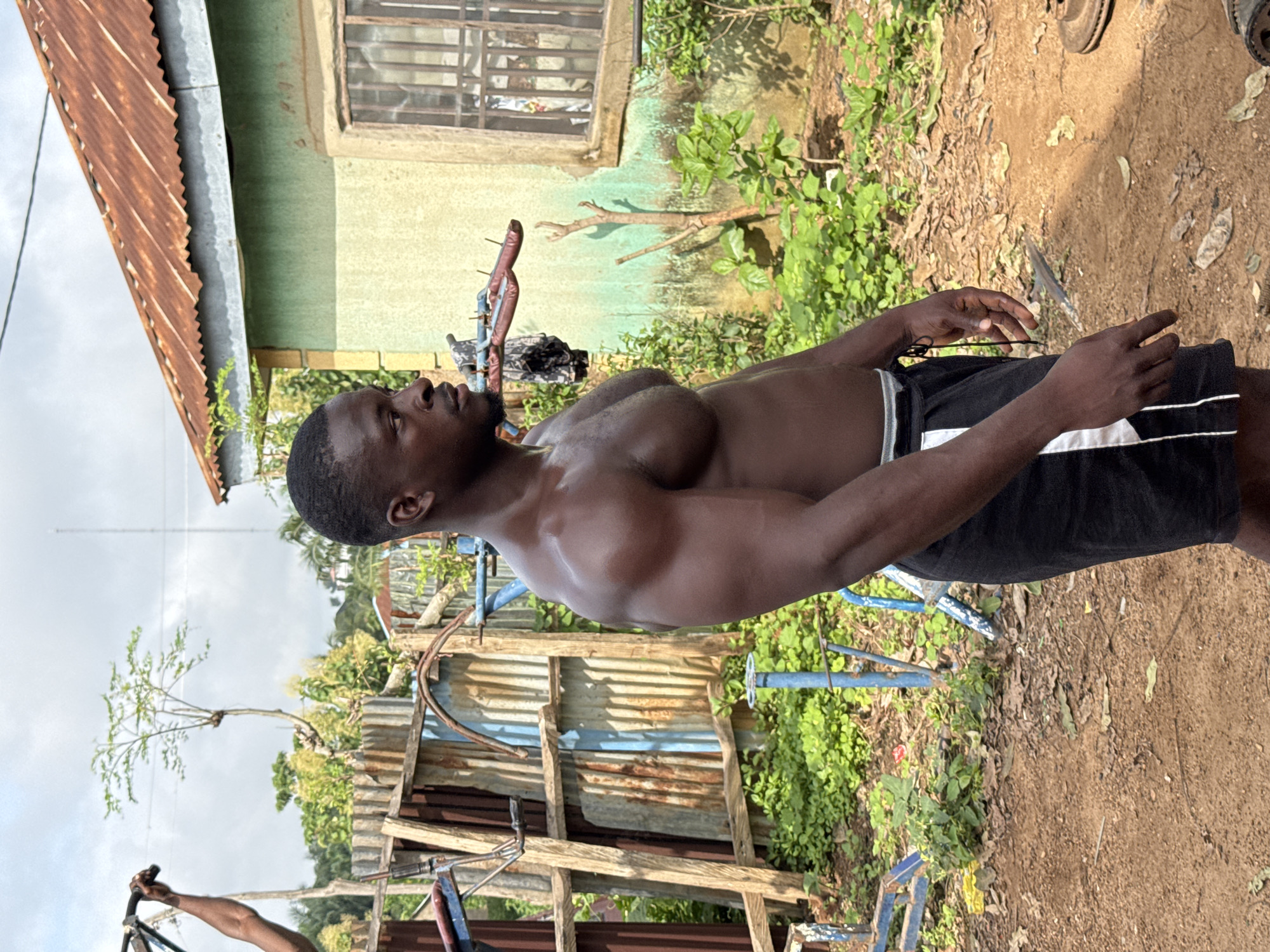 A muscular Black man is standing outdoors, facing right in profile. He has dark skin and is shirtless, wearing black athletic shorts with white stripes down the sides. His physique is well-defined, particularly his shoulders and chest. He appears to be in an outdoor gym or workout area, suggested by the presence of exercise equipment, including a blue metal structure with bars behind him. The environment is somewhat rustic, with corrugated metal fencing and wooden structures visible, along with lush green vegetation and dirt ground. In the background, a light green building with a rusty metal roof and a barred window is situated. The sky is partly cloudy, with a tall radio tower visible in the distance. The scene is set in Akwanga, Nigeria, indicated by the context provided.