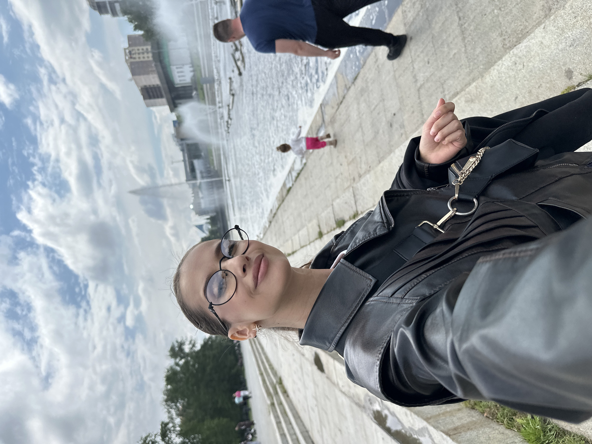 A young woman with dark hair and glasses takes a selfie while standing on a paved embankment. She is wearing a black leather jacket. Behind her, a large fountain is active, with water spraying upwards. In the middle ground, a small child in pink pants and a white shirt runs towards the fountain, and a man in a blue t-shirt and black pants walks away from the camera. The background features several modern buildings and trees. The scene is set in Yekaterinburg, Russia, likely near the Iset River.