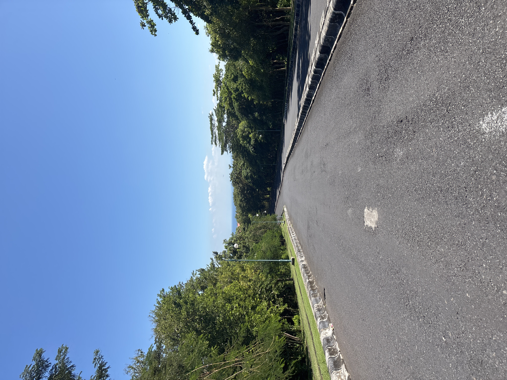 The image shows a paved road with a decorative curb running along one side, lined with green grass and trees. Streetlights are visible among the trees. The clear blue sky is on the left of the frame. A glimpse of what appears to be the ocean can be seen in the distance through a break in the trees. Based on the location, Pecatu, Indonesia, the road is likely near the coastline. There are no people visible.