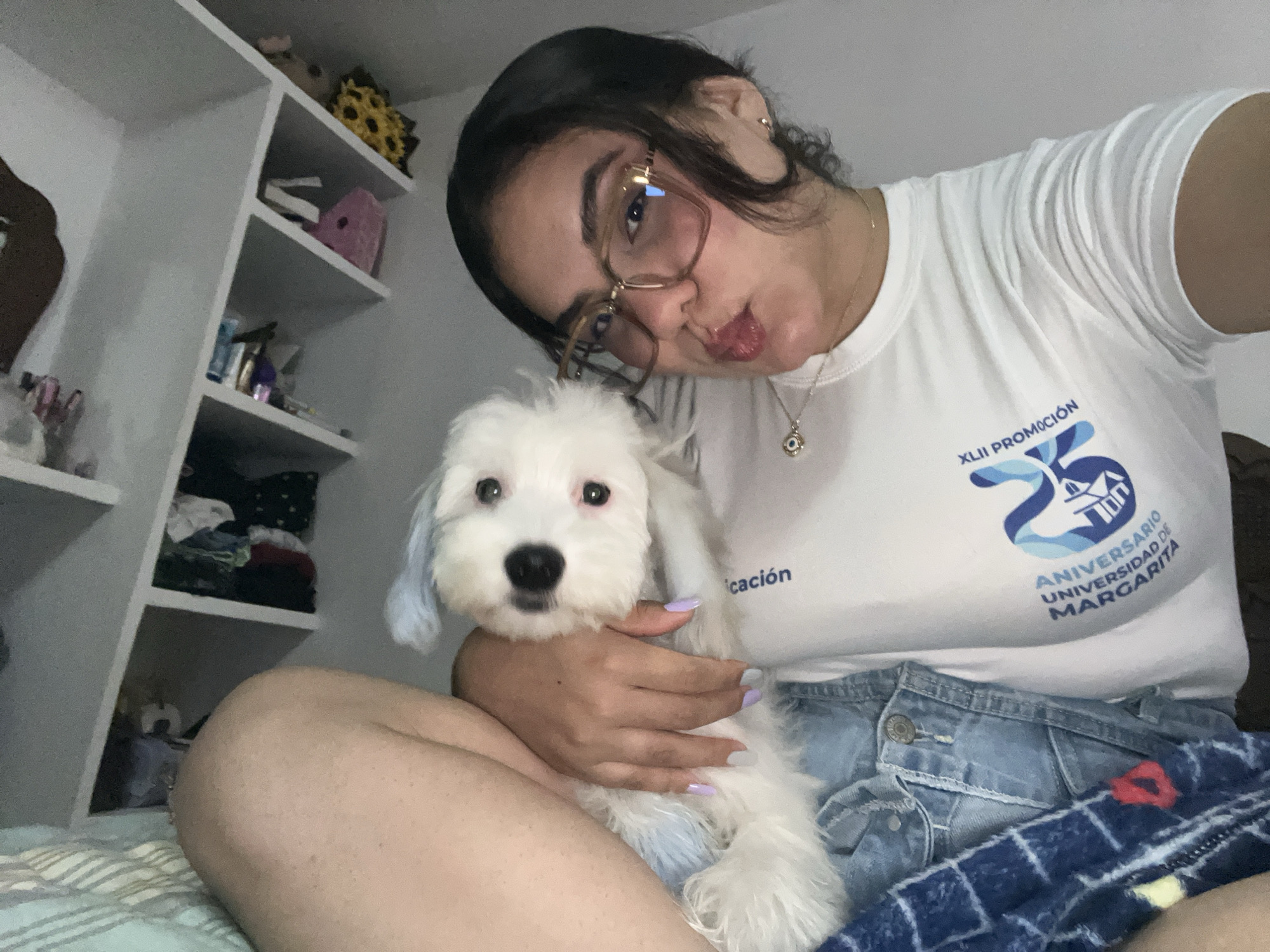 A young woman with dark hair and glasses, wearing a white t-shirt and denim shorts, is taking a selfie with her small white dog. The dog, which has light blue tips on its ears, is being held by the woman. They are seated on a bed with a striped blanket, in front of shelves filled with various items. The t-shirt has a blue logo with text that appears to be related to an anniversary, possibly "ANIVERSARIO UNIVERSIDAD DE MARGARITA". The environment suggests an indoor setting, likely a bedroom. The scene is intimate and casual, capturing a personal moment.