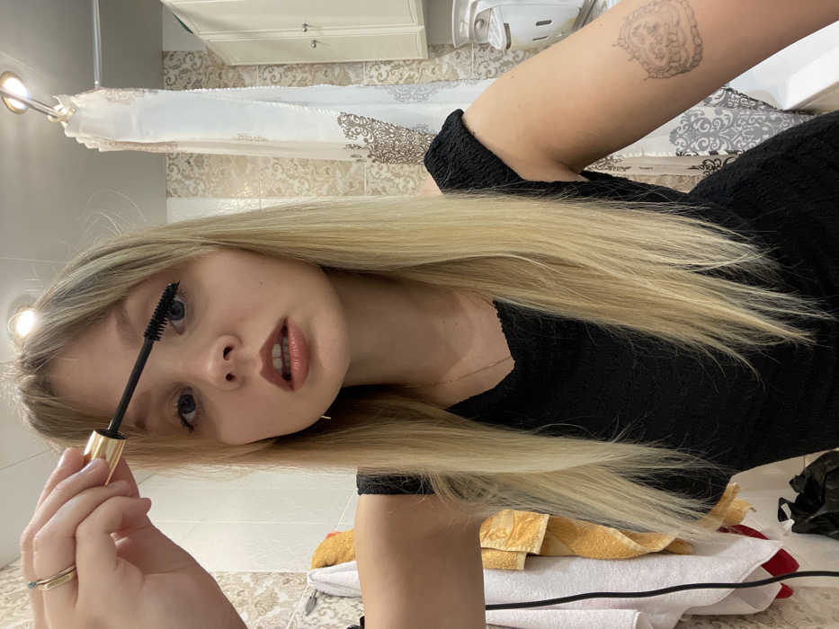 A young Caucasian female with long blonde hair and bangs is depicted in a bathroom, applying mascara to her right eye using a black-bristled wand with a gold handle held in her left hand. She wears a black textured top, a gold ring with a green stone on her left ring finger, a thin gold necklace, and a small gold earring on her right ear. An ornate, light-colored, heart-shaped tattoo is visible on her right upper arm. The background features light-toned patterned wall tiles, a yellow and white towel hanging on the left, a partially visible white shower curtain with a brown pattern on the right, and a white two-door cabinet in the upper right. A crumpled black plastic bag is present on the lower left. Two overhead light fixtures illuminate the scene. This scene occurs in a bathroom in the city of Vysokovo, Russia.