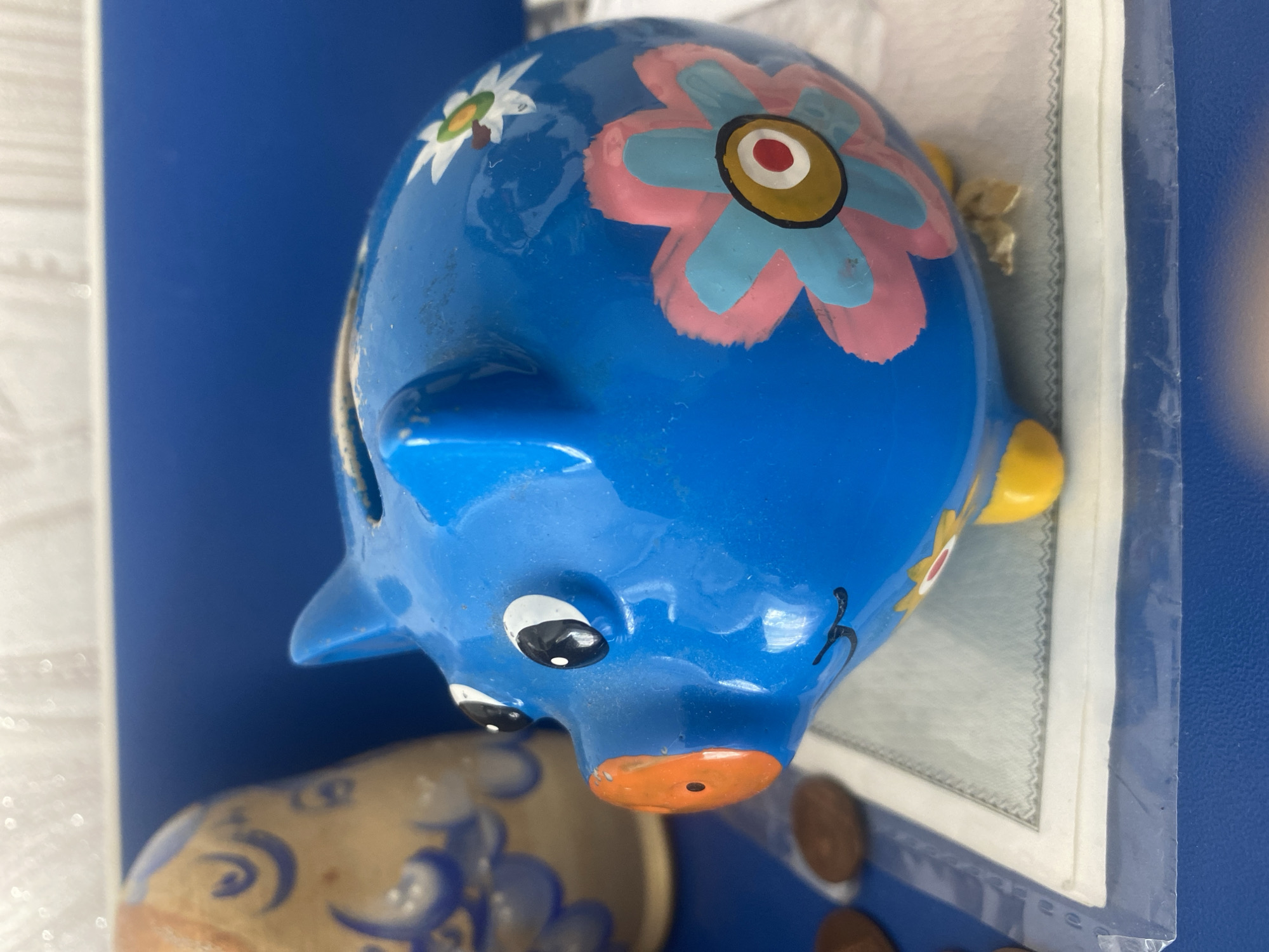 A blue ceramic piggy bank painted with pink and blue flowers sits on a white surface with a patterned border, partially covered by clear plastic. The piggy bank is shaped like a pig, with painted black eyes, a smiling mouth, and an orange snout. Yellow feet support the body. To the left of the piggy bank is a portion of a wooden object decorated with blue patterns, possibly a matryoshka doll. A few brown coins are visible in the lower left corner of the image. The background consists of a blue surface and a textured white wall. The scene is brightly lit, with some specular highlights visible on the piggy bank's surface. There are no people or animals present. The image does not provide any information about its location in Vitsebsk, Belarus.