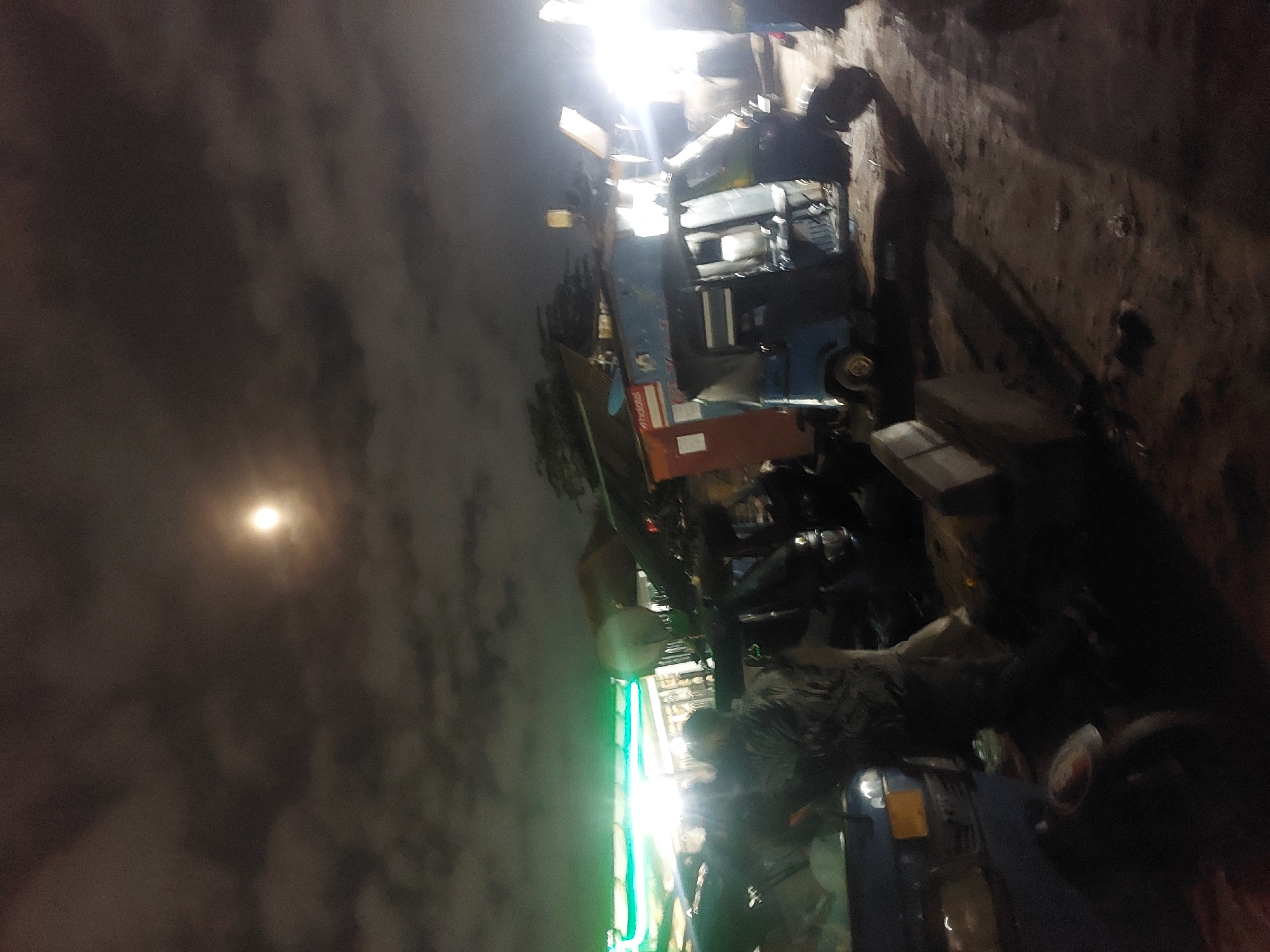 The image depicts a street scene at night in Dar es-Salaam, Tanzania. The image is dark, with limited visibility. The left side of the frame shows the night sky with the moon visible through clouds.
The right side of the image shows a rough ground surface with shadows and debris. The scene is illuminated by a mix of artificial lighting sources, including bright overhead lights and a vertical green light source.
The visible objects include a vehicle, possibly a type of public transport such as a minibus or daladala. A possible storefront with green light could be visible near the street. There may be people visible in the crowd, but the dark and grainy quality makes details hard to ascertain.
