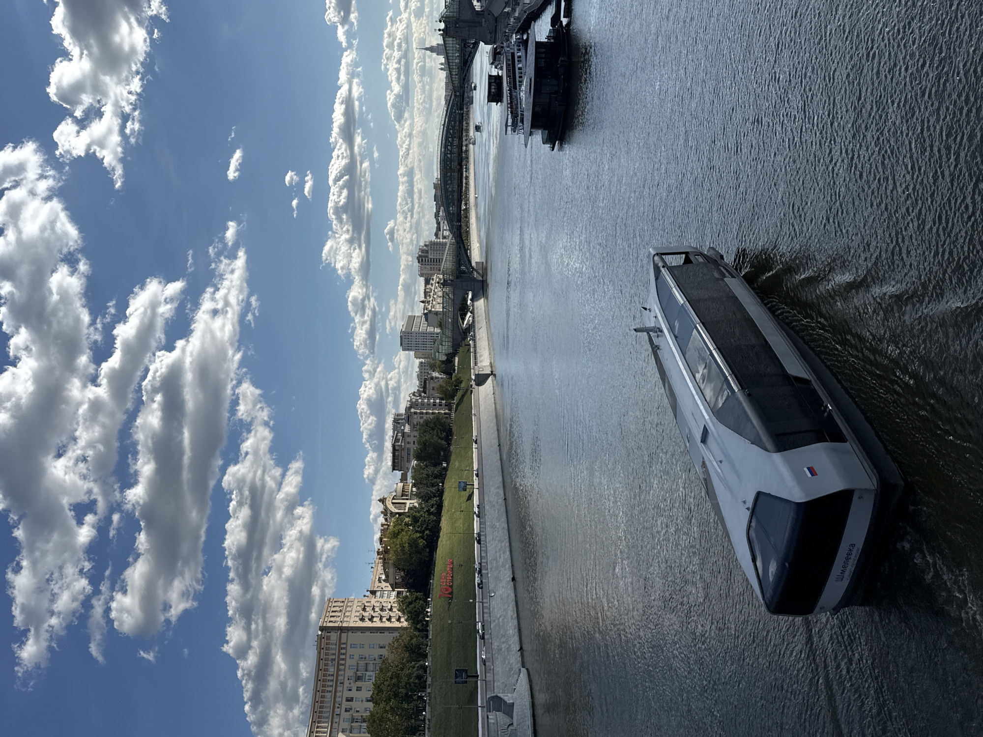 An elevated, eye-level, medium shot captures a wide river in Moscow, Russia, on a clear day with scattered white clouds. In the foreground, a sleek, modern, grey and black passenger boat, named "Шмелёвка" (Shmelevka) according to visible text on its stern, moves through the water. Its bow is angled toward the left of the frame, with a wake trailing behind it. The river itself is a dark, rippled expanse, reflecting the sky.
Along the left bank of the river, a concrete embankment leads up to a grassy slope. On the slope, large red letters spell out "70 ДЕНЬ СТРОИТЕЛЯ" (70th Builder's Day). Behind the slope, a multi-story, tan-colored apartment building with numerous windows dominates the skyline. Further down the bank, trees and other buildings are visible.
Across the river, a modern, multi-span pedestrian bridge with a glass enclosure extends across the water. Several other boats are moored or cruising in the distance, including a larger, dark-colored vessel tied to a pier on the right bank. The cityscape in the background features a mix of architectural styles, including modern high-rise buildings and older structures. The overall scene depicts a vibrant urban waterway with a blend of historic and contemporary elements.