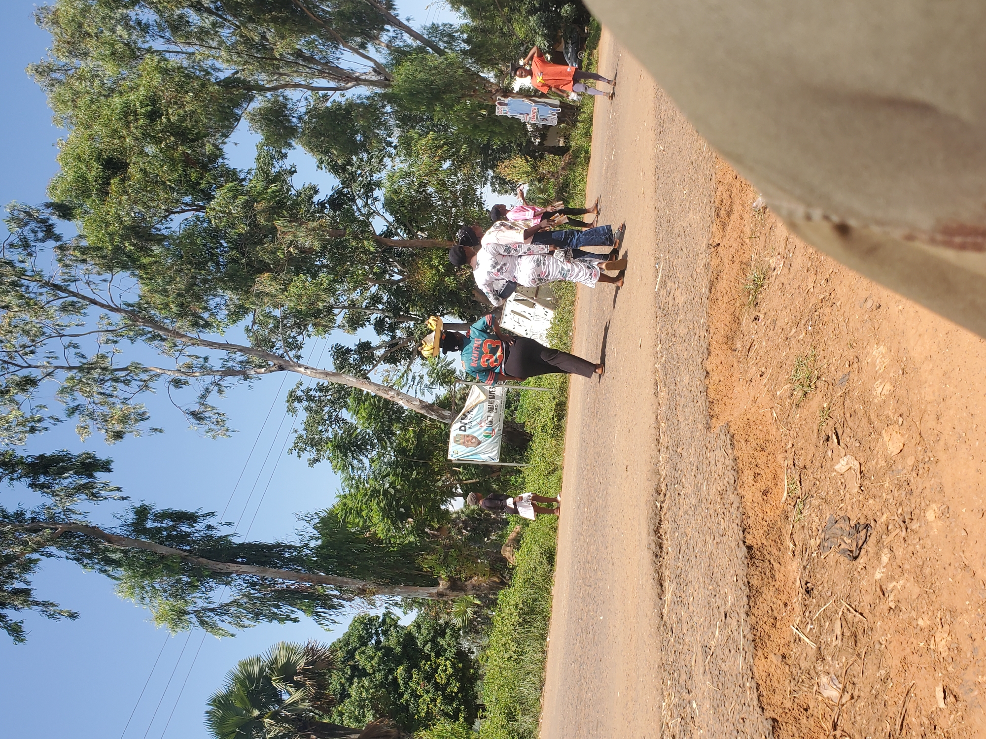 In Mangu, Nigeria, a group of people are seen walking along a dirt road. The scene is set under a clear blue sky, suggesting a sunny day. Lush green trees and vegetation line the side of the road, with some of the trees appearing to be eucalyptus.
The people are dressed in various casual clothing. One individual in the foreground wears a blue top with the number '23' on it and a black skirt, carrying a tray of items on their head. Others are dressed in colorful patterned outfits. A banner is visible in the background with a picture of a man's face and text that appears to be related to an election or campaign, with "SENATE" being partially legible. Another person in an orange shirt stands further down the road, holding a cutout resembling a campaign sign.
The overall impression is of a lively, possibly communal gathering or a journey taking place on a warm day in a rural Nigerian setting. The people appear to be walking with purpose, and the presence of the banner suggests a local event or political activity. The image is captured in portrait orientation.