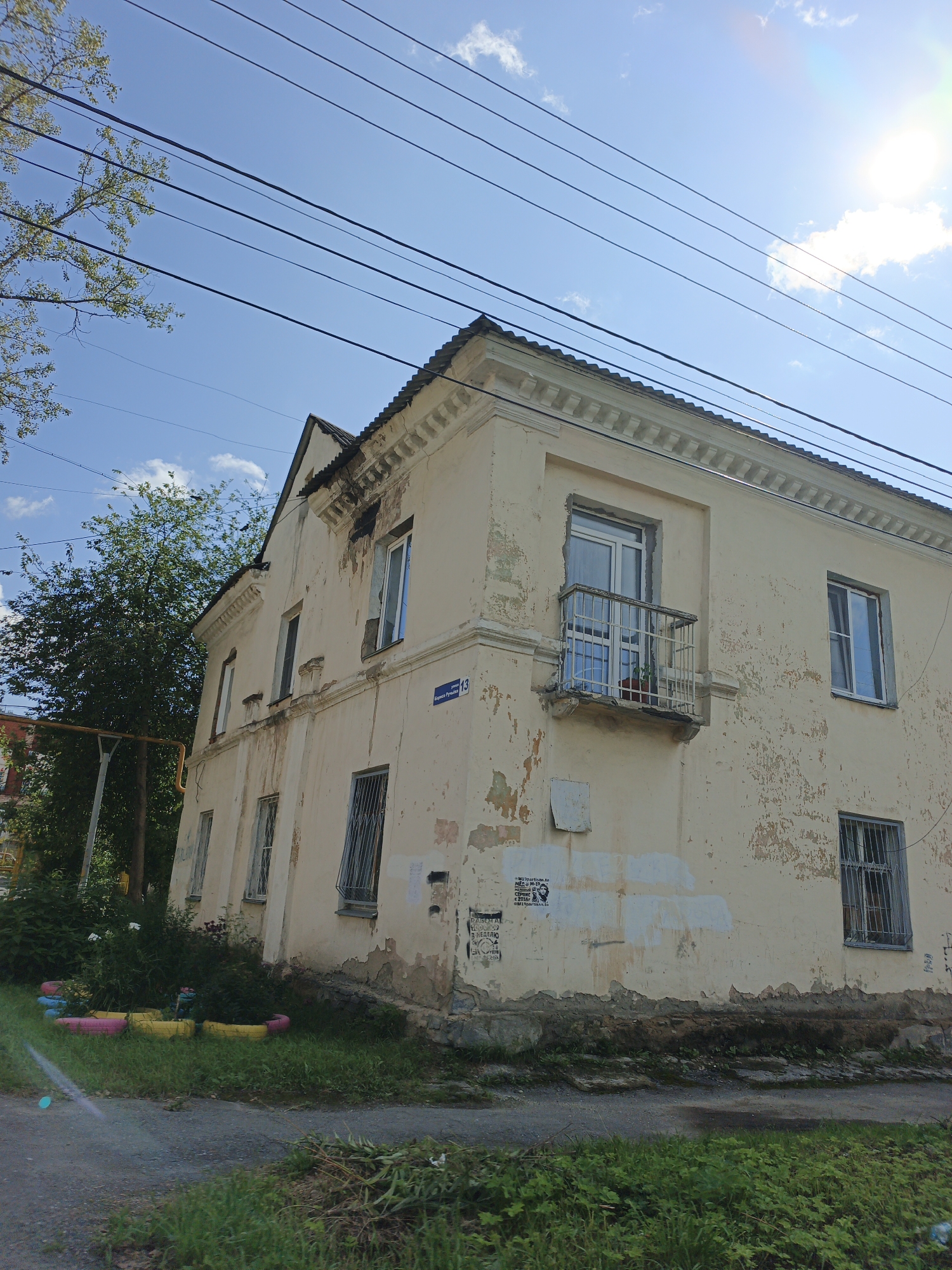 The image depicts a weathered, two-story apartment building with a light yellow facade, located in Zlatoust, Russia. The building shows signs of disrepair, with peeling paint and crumbling plaster. There are several windows on both floors, some with metal grates. A small balcony with a white railing is present on the second floor. Overhead, multiple power lines stretch across the bright blue sky, which is partially obscured by a bright sun and a few scattered clouds. To the left of the building, a large, leafy green tree stands tall. In front of the building, a patch of grass is visible, with some colorful repurposed tire planters in the foreground. A gray asphalt path runs along the bottom of the image, with more greenery and some scattered debris. There are no people visible in the scene.
