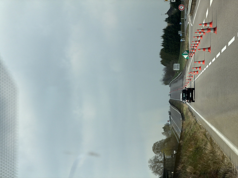The image depicts a multi-lane road scene in Pagny-sur-Meuse, France, viewed from inside a vehicle's front passenger compartment. The sky is overcast with a light grey cloud cover. The primary subject is a paved asphalt road extending into the distance, ascending a gentle incline.
In the foreground and middle ground, the road features white dashed and solid lane markings. Directly ahead, in the right lane, a dark-colored Dacia Duster SUV is proceeding forward. To the right of the Duster, a long line of orange and white traffic cones marks a temporary lane closure or redirection, guiding traffic towards the right. This cone-delineated path leads to an intersection or diverging road branch, where a circular white speed limit sign indicating "70" is visible on the right shoulder, alongside a green diamond-shaped sign with white arrows indicating a split in directions.
On the left side of the main road, a grassy embankment borders a separate, elevated road or overpass that also ascends into the distance. Several indistinct vehicles are visible on this upper road and in the far distance on the main thoroughfare. The immediate roadside on the left features a concrete barrier and sparse, deciduous trees. On the right, beyond the coned-off section, there are clusters of evergreen trees and a partial view of a building with a dark roof. No individuals are visible in the scene. The overall environment suggests a rural or semi-rural highway context with ongoing road adjustments.