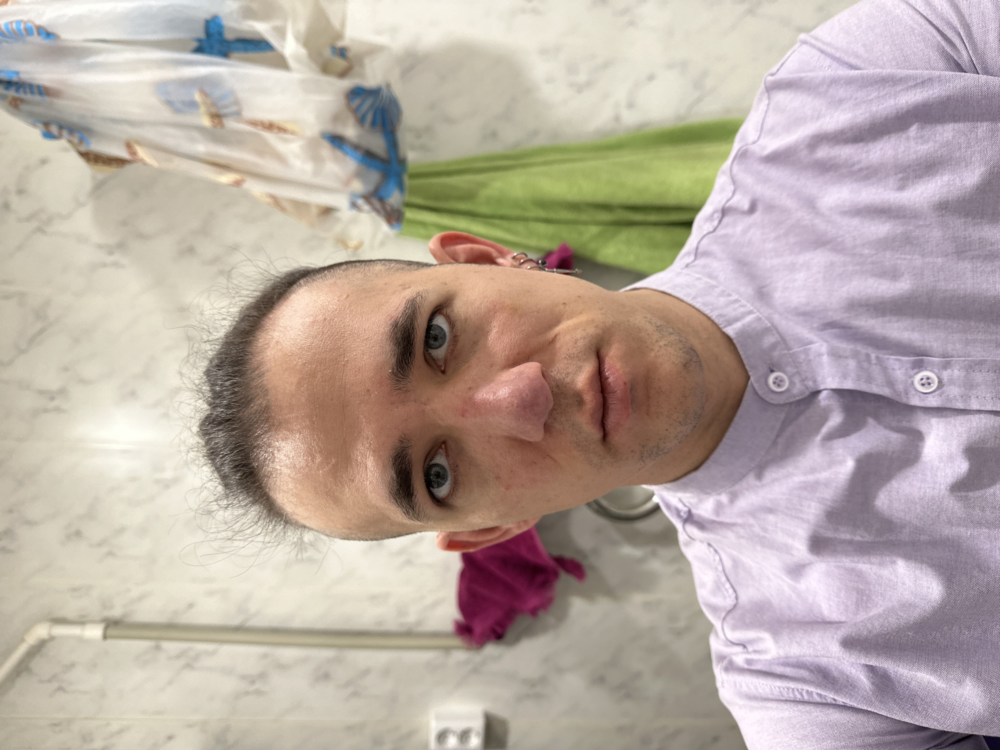 A selfie of a person with closely cropped dark hair, blue eyes, and fair skin wearing a lavender button-down shirt is taken indoors. The person is looking at the camera. Behind the person is a bathroom setting with a marble-patterned wall, a towel rack, a white towel with blue and tan patterns, a green towel, and a pink towel. There is an electrical outlet near the bottom of the frame. The person has multiple piercings in the left ear. There is no indication in the image that the location is Saratov, Russia.
